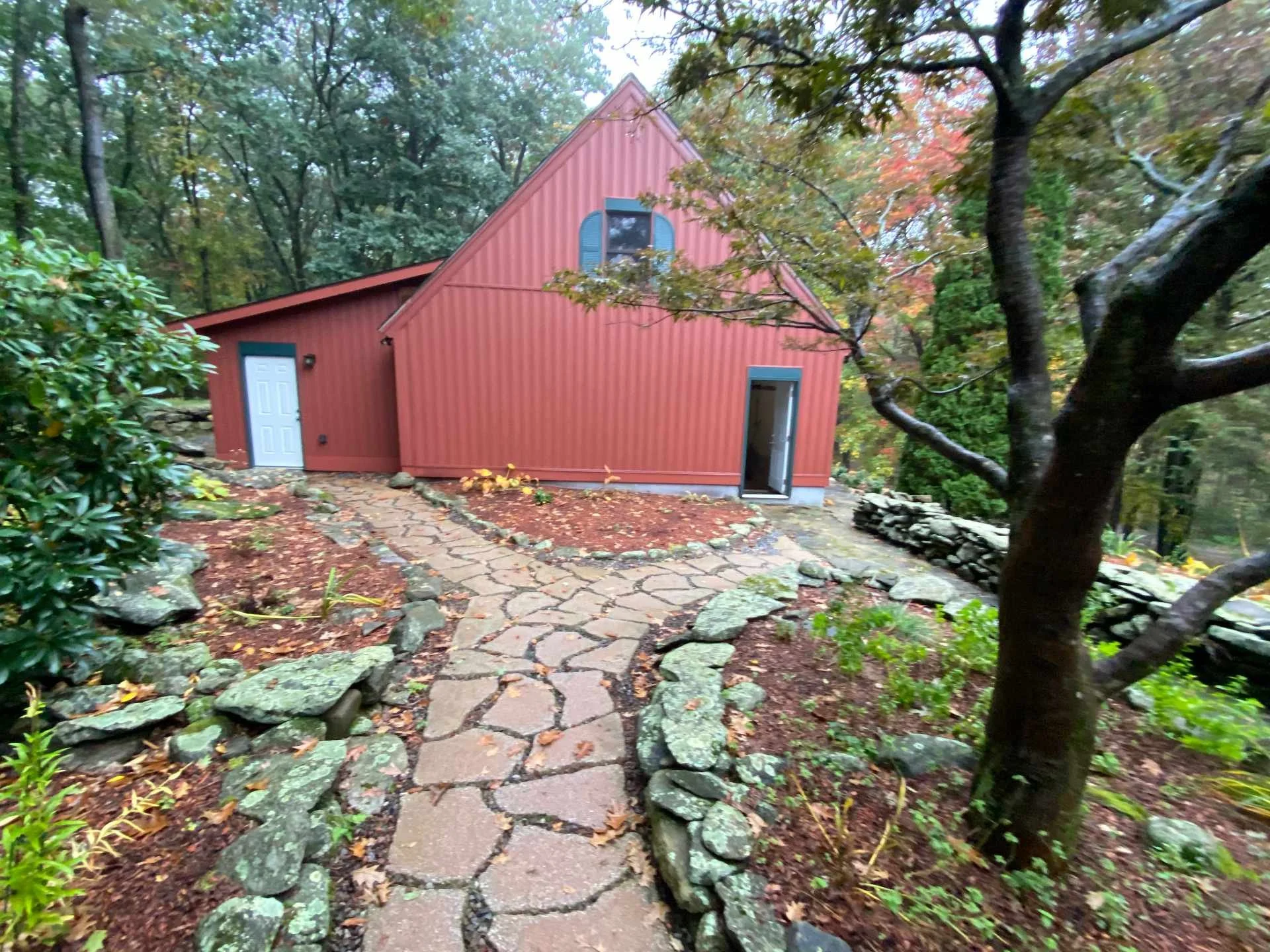 A red house with a steep roof surrounded by trees with fall foliage, a stone pathway leading to the house, and a small white door.