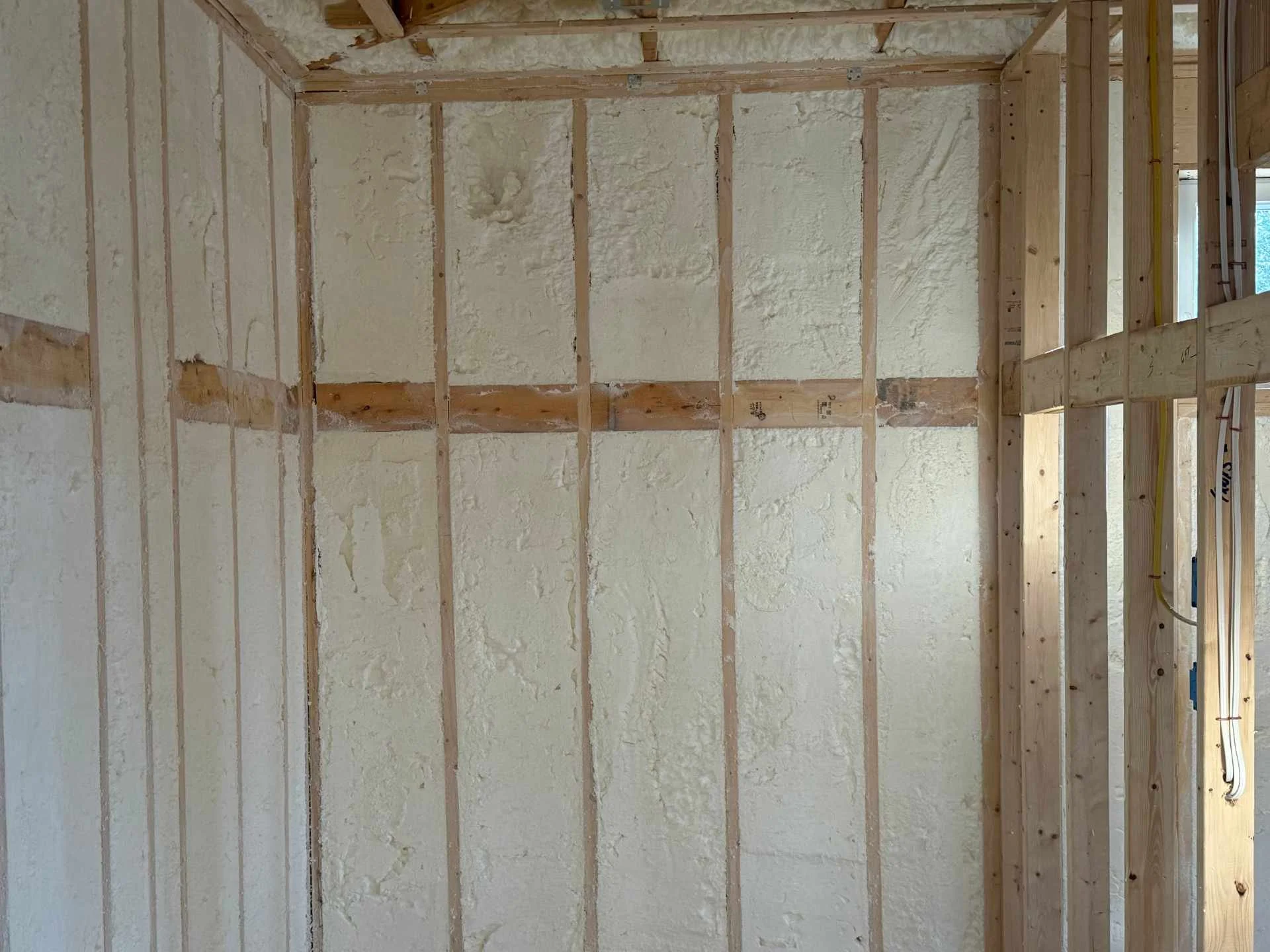 Interior view of a building under construction with exposed wooden framing and spray foam insulation in the walls.