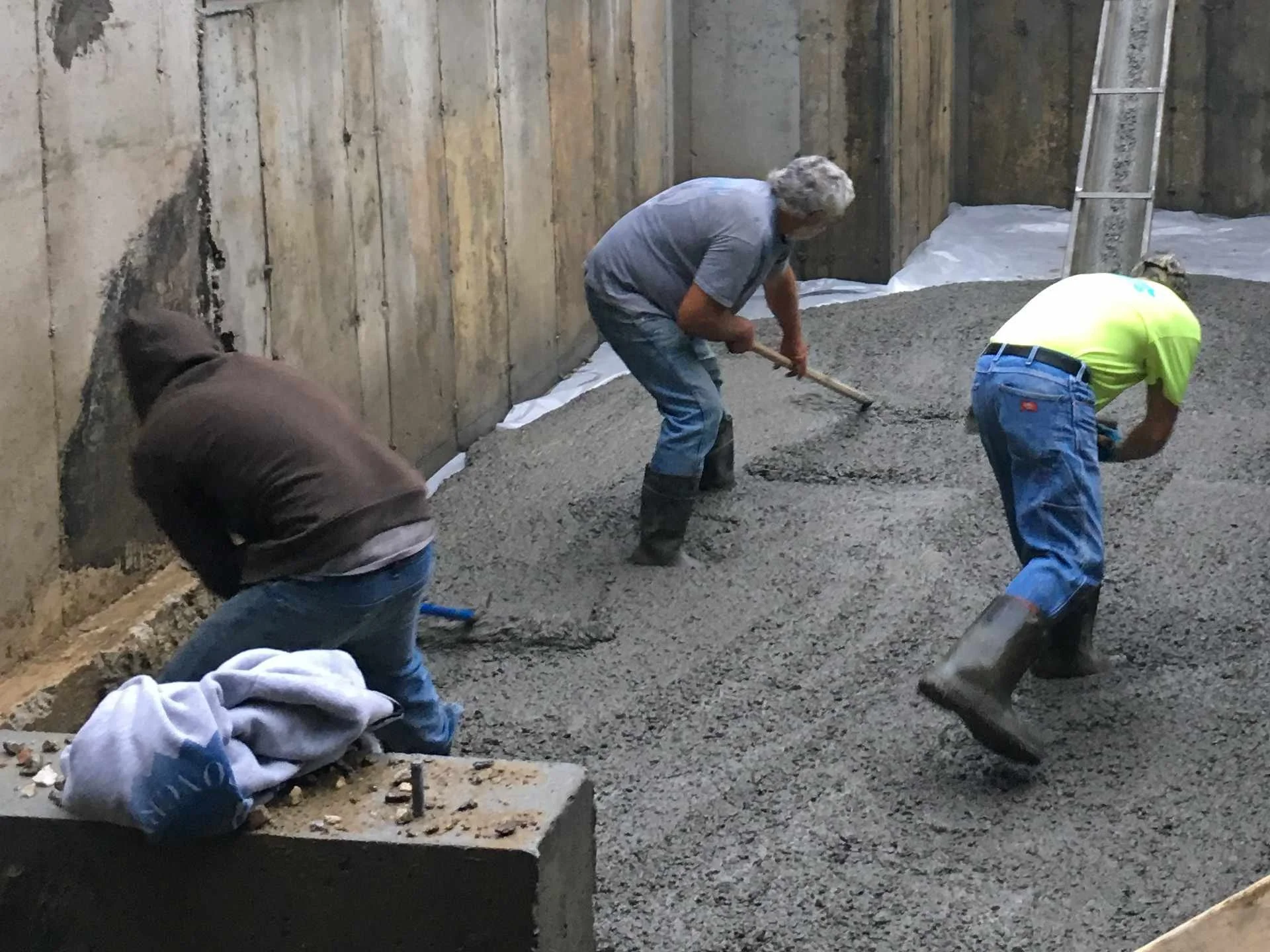 Three workers, one with gray hair, are backfilling concrete in a construction site with a wooden wall on the left and a ladder on the right.
