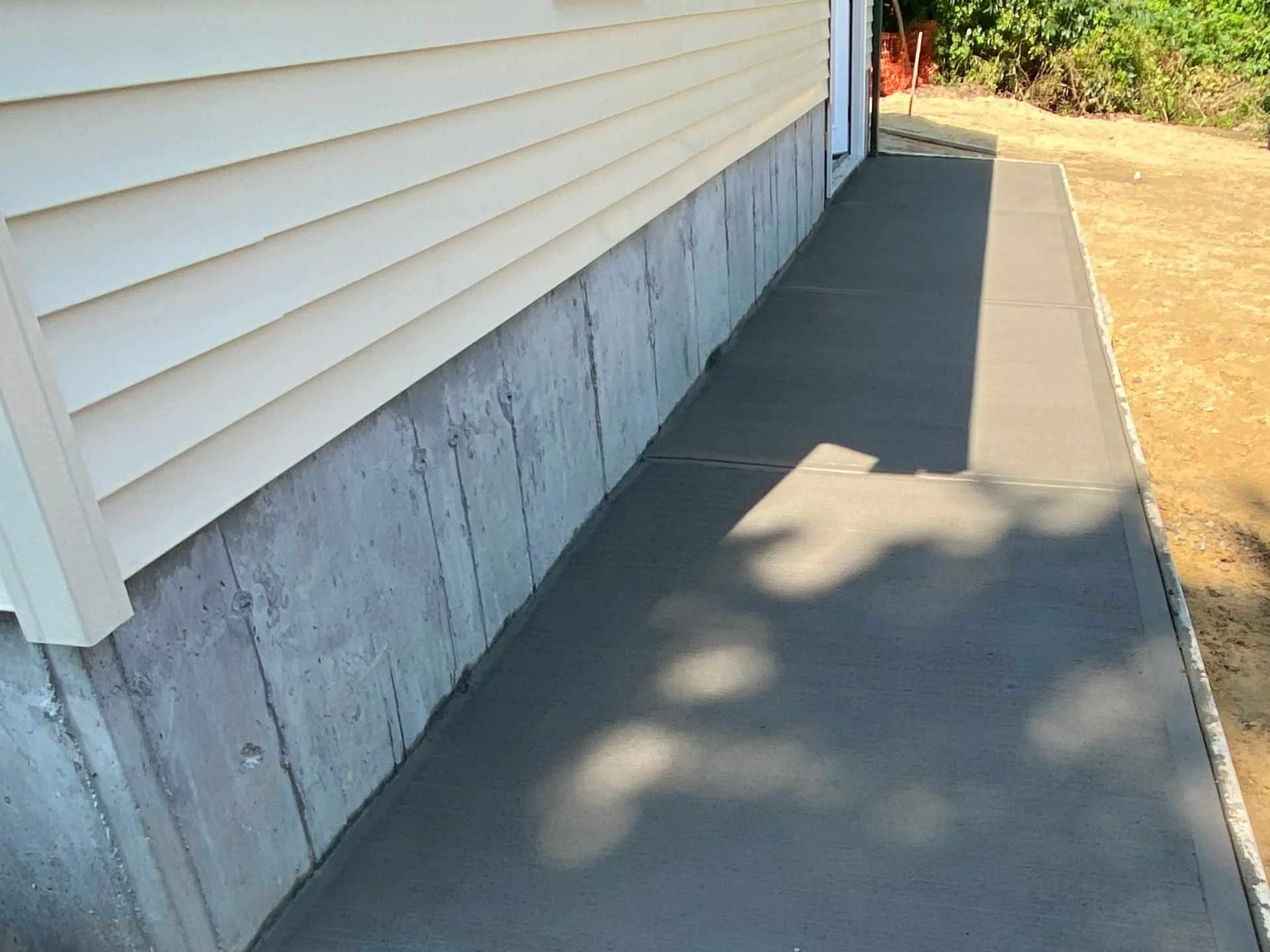 A concrete sidewalk outside a house with beige vinyl siding and a concrete foundation. There is a shadow cast on the sidewalk, and some greenery and dirt are visible at the end of the sidewalk.