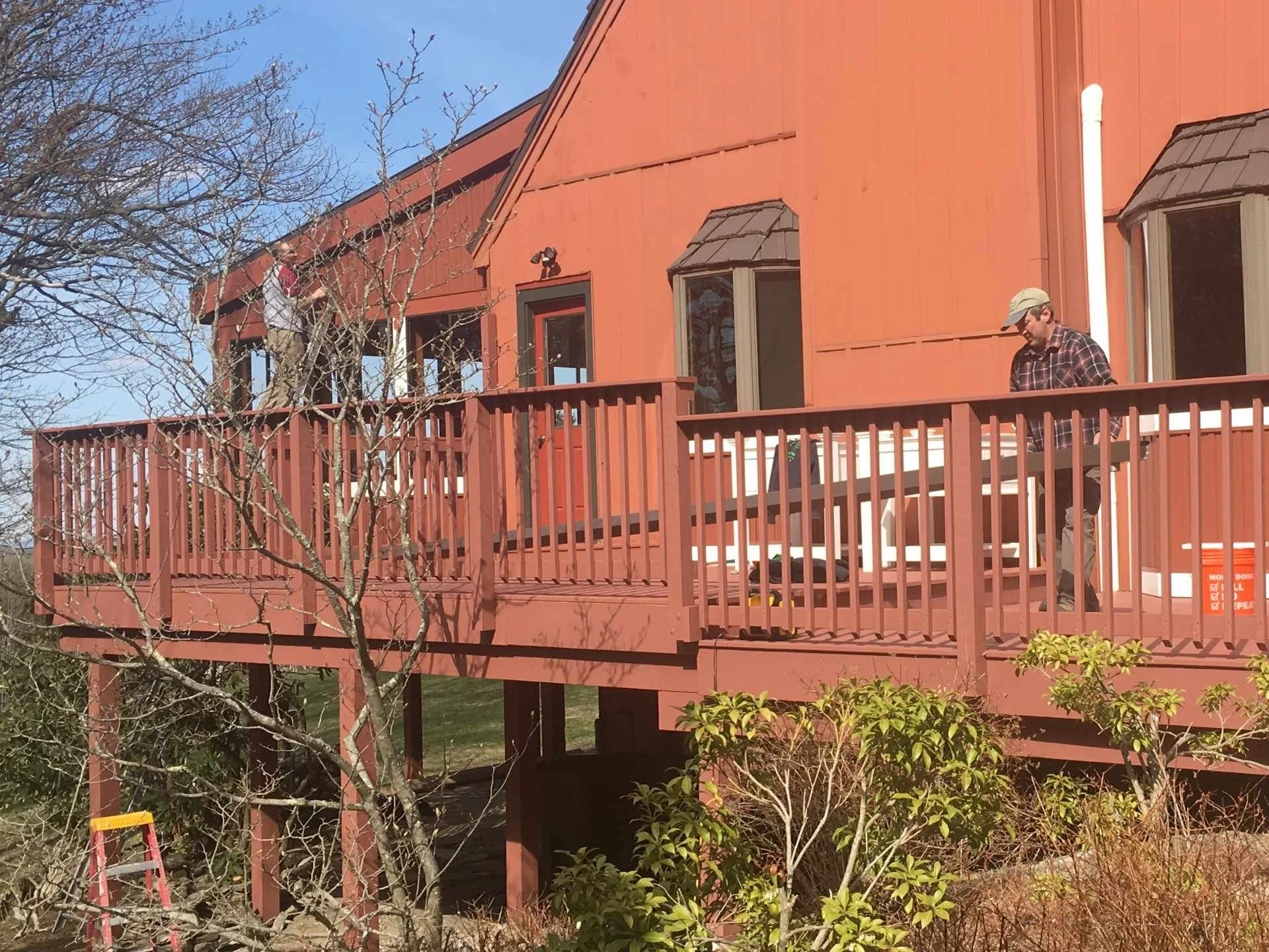 A two-story deck with a glass door and windows, under construction, with people working on it, surrounded by trees and bushes, and a clear blue sky.