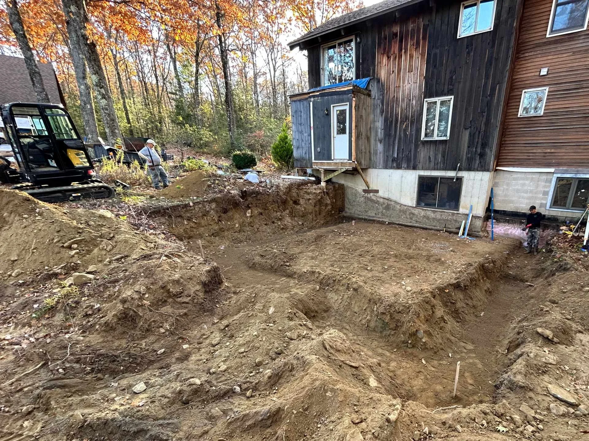 Excavation site next to a house with trees in the background, showing workers and construction equipment preparing for foundation work.