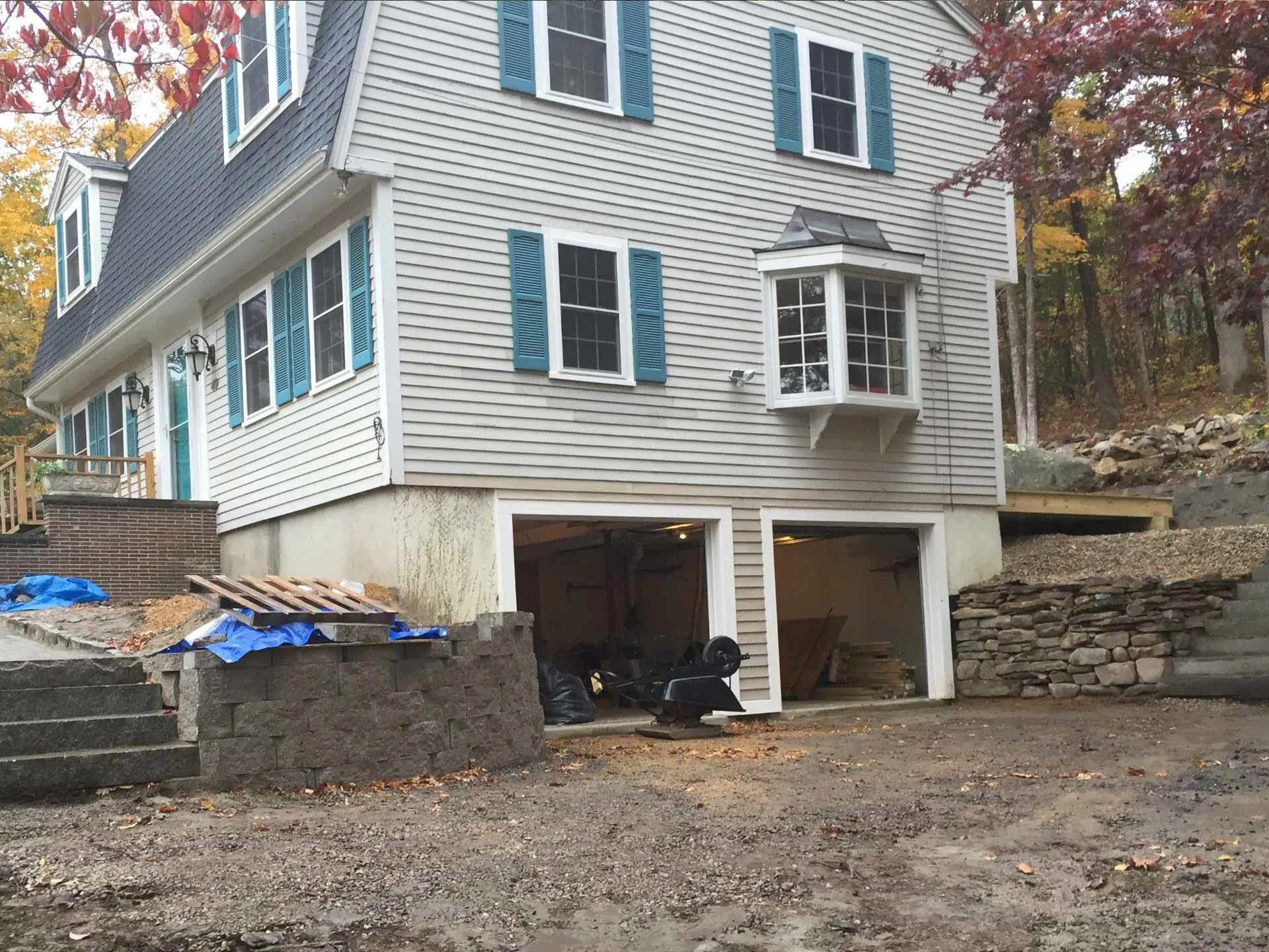 The image shows a multi-story house under construction or renovation, with the garage area open and unfinished. The house has beige siding with blue shutters on the windows. There are trees with fall foliage in the background. Construction materials 