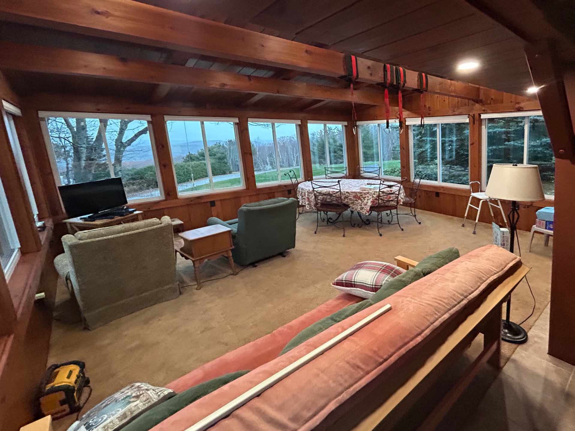 A living room with large windows showing trees and a landscape outside. The room has wooden walls and ceiling, a sofa, chairs, a TV, a table with a floral tablecloth, a lamp, and assorted furniture and items, indicating ongoing home improvement or re