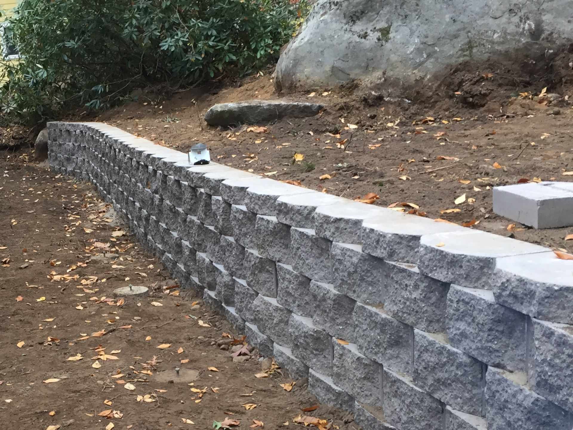 A curved stone engineered retaining wall with a lantern on top, fallen leaves on the ground, and bushes and rocks in the background.