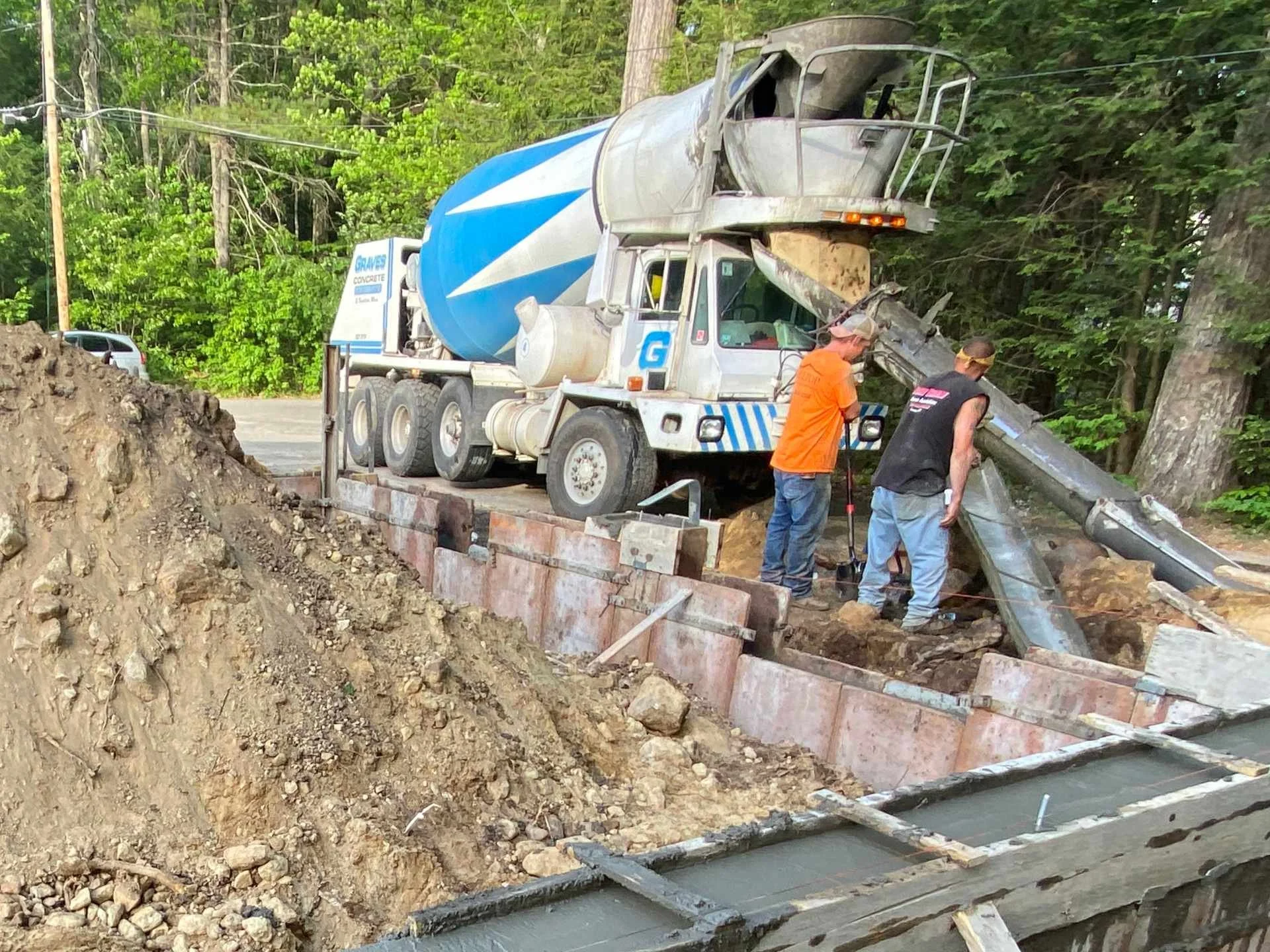 A cement truck pouring concrete into formed foundation walls on a construction site, with workers standing nearby and green trees in the background.