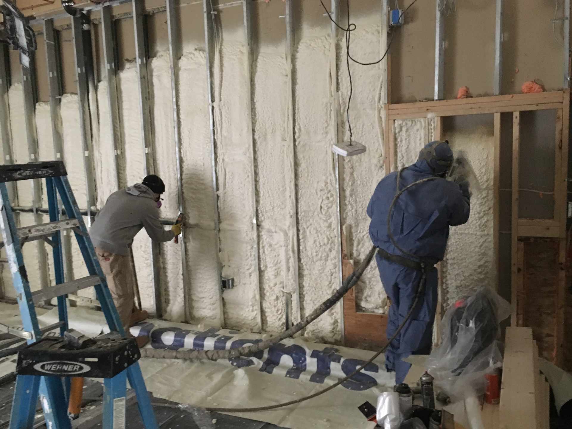 Two workers applying spray foam insulation to a wall in a construction site, with ladders and tools nearby.