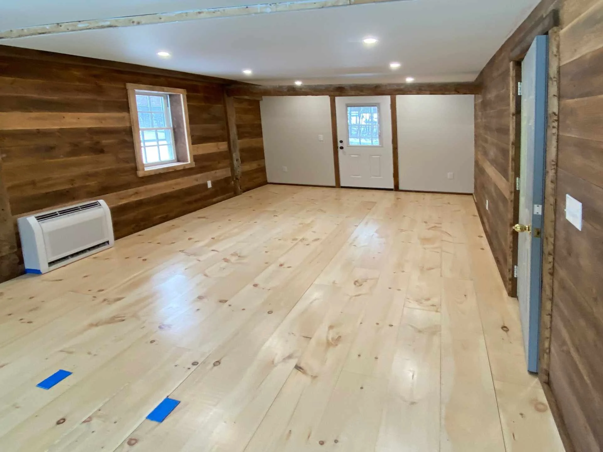 Home office room built inside existing post and beam barn with wooden walls and flooring, two small windows, a door, and Fujitsu floor-mounted mini-split heating/cooling unit