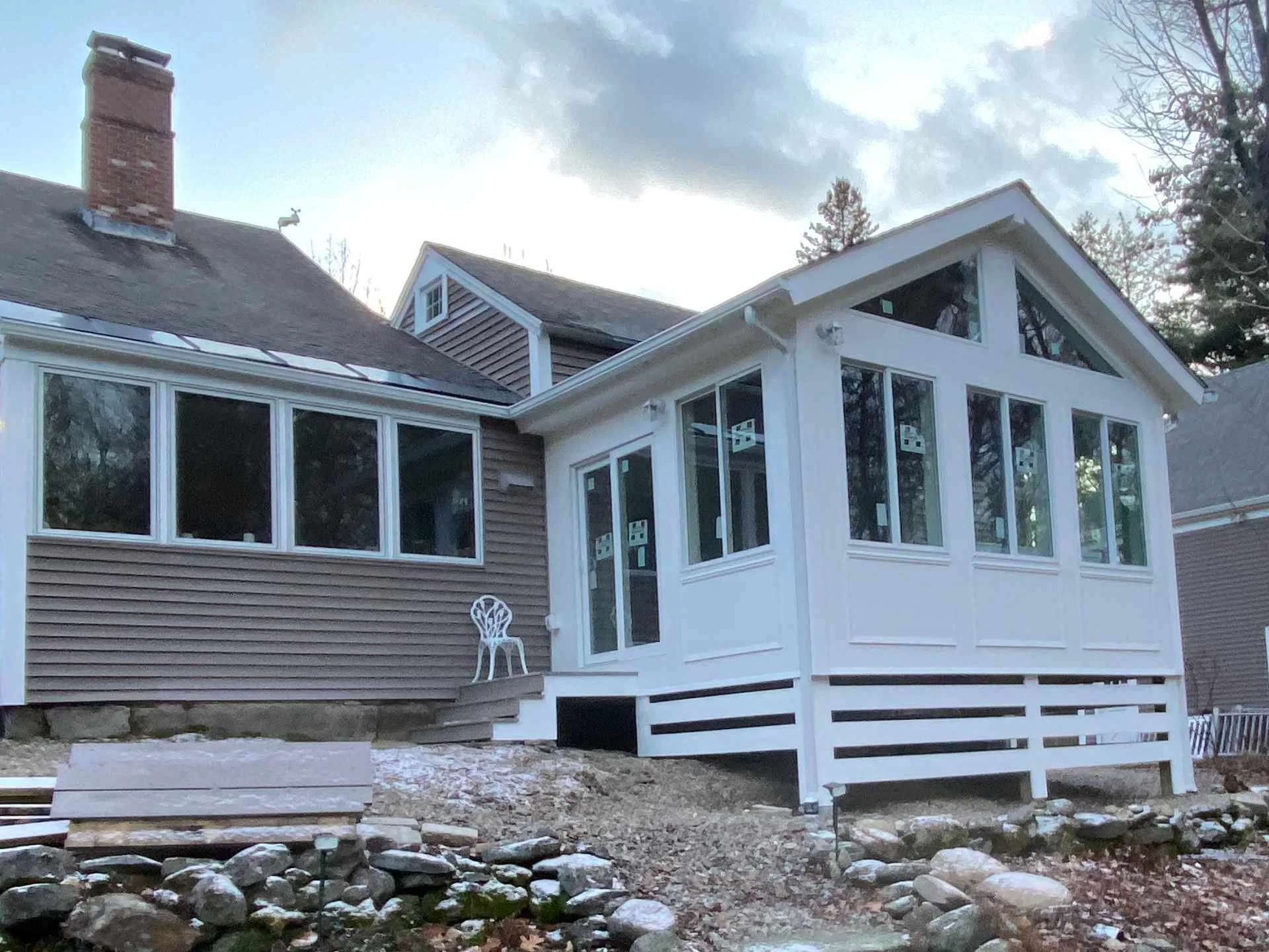 The image shows a house with a modern extension featuring large windows and a white framework. There is a small white chair on the ground outside the house. The ground appears rocky and uneven, with some patches of dirt. The sky is partly cloudy, and