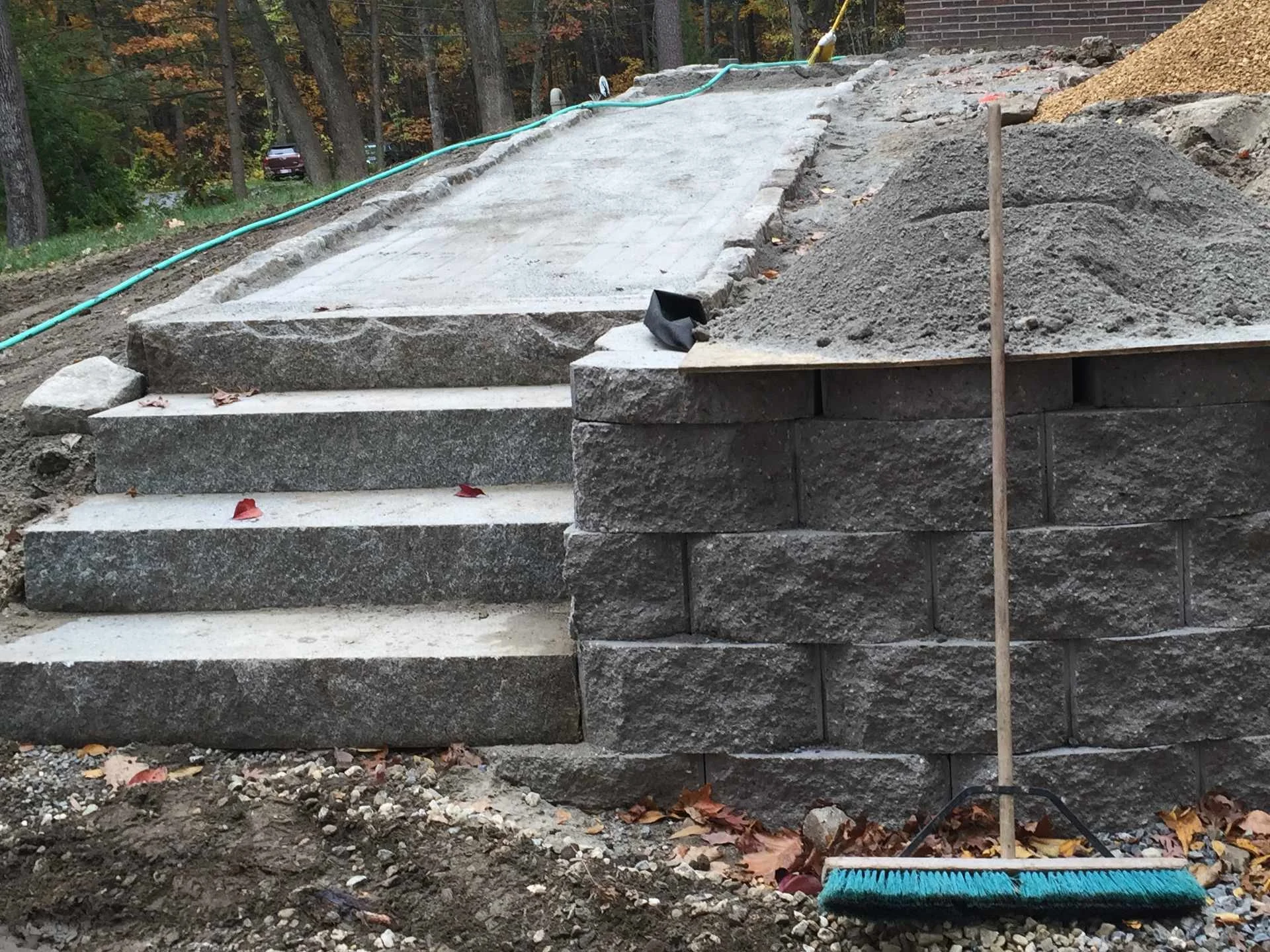 Stone steps under construction surrounded by dirt, with a garden rake in the foreground and a hill of gravel on the right.