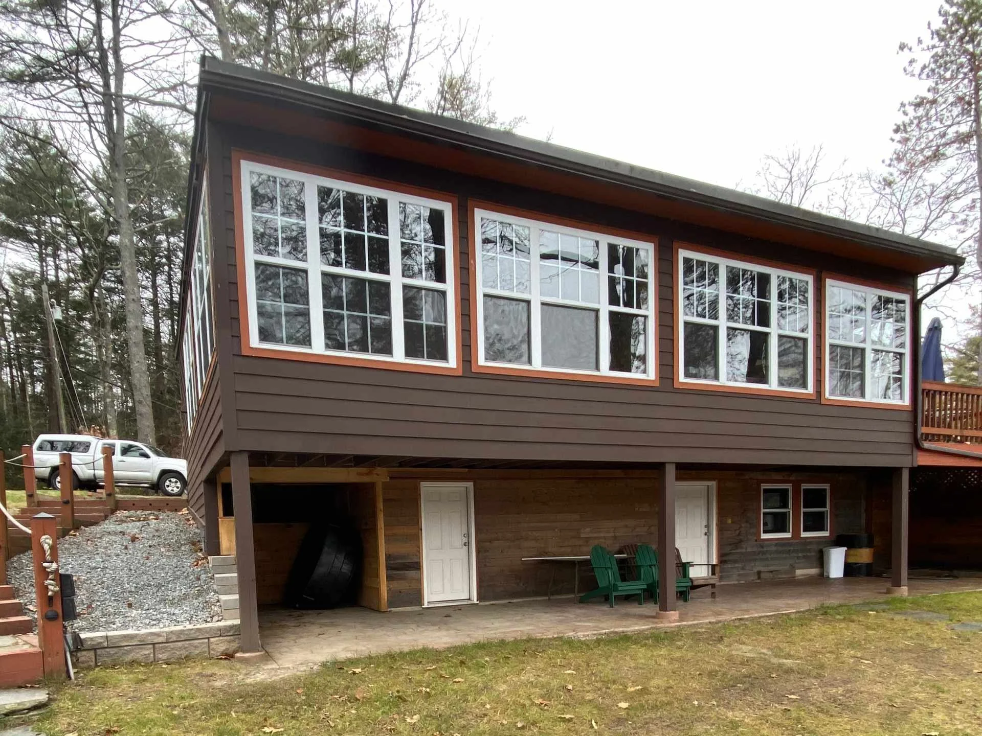 A two-story house with brown siding and large front windows, surrounded by trees with no leaves, indicating fall or winter. The house has a covered area on the ground floor with two white doors, and outdoor furniture including green chairs and a smal