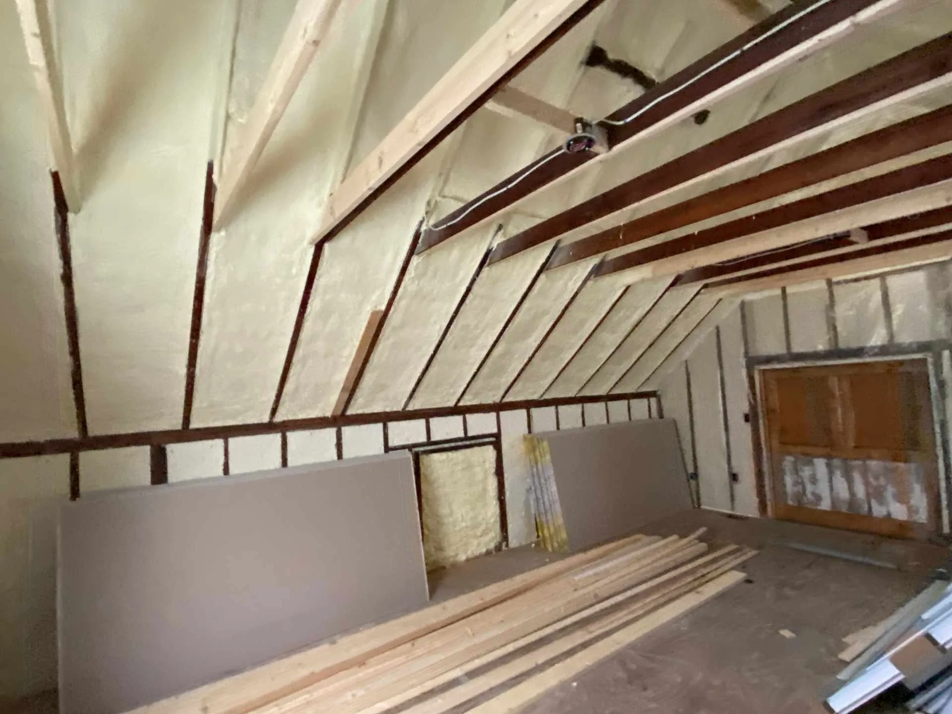 Interior of a room under construction with exposed wooden ceiling beams and spray foam insulation, construction materials and tools are visible.