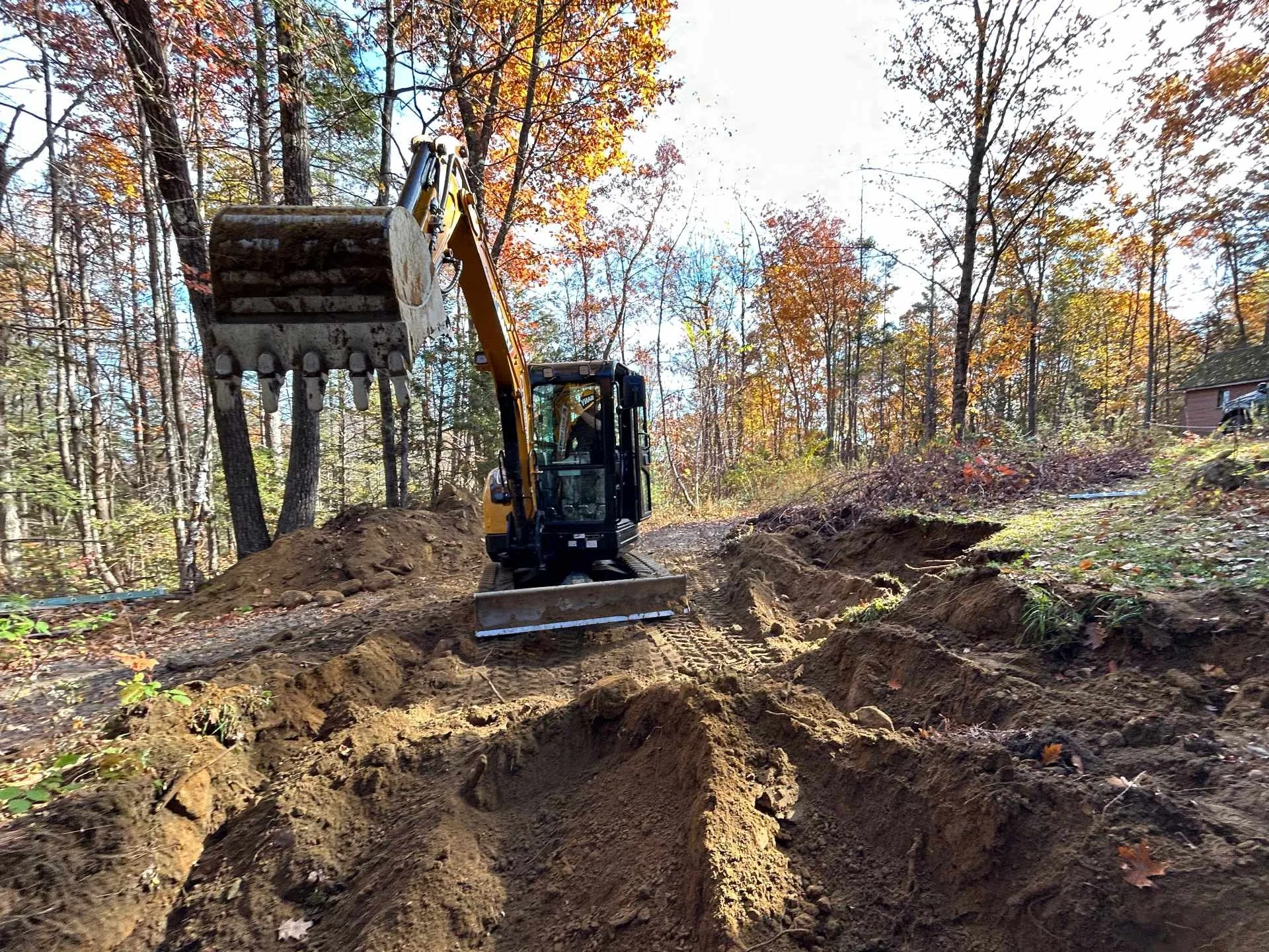 A construction excavator working in a wooded area during autumn, digging up soil with trees and a small building in the background.