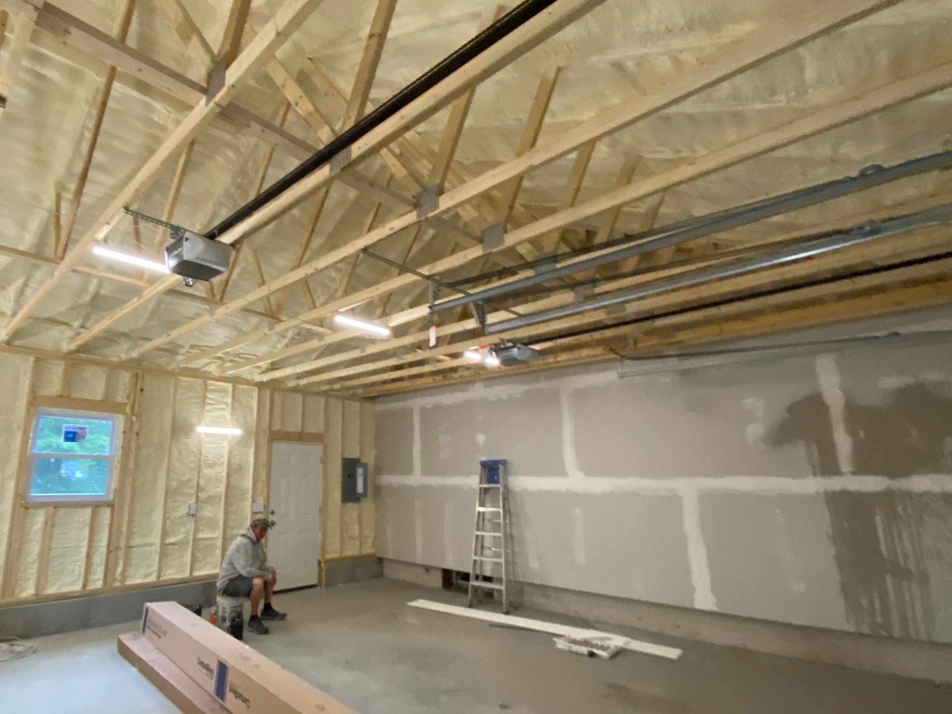 Interior view of a partially constructed garage or room showing wooden framing, spray foam insulation, and some construction materials. A man is sitting on a box, and a ladder is against the wall.