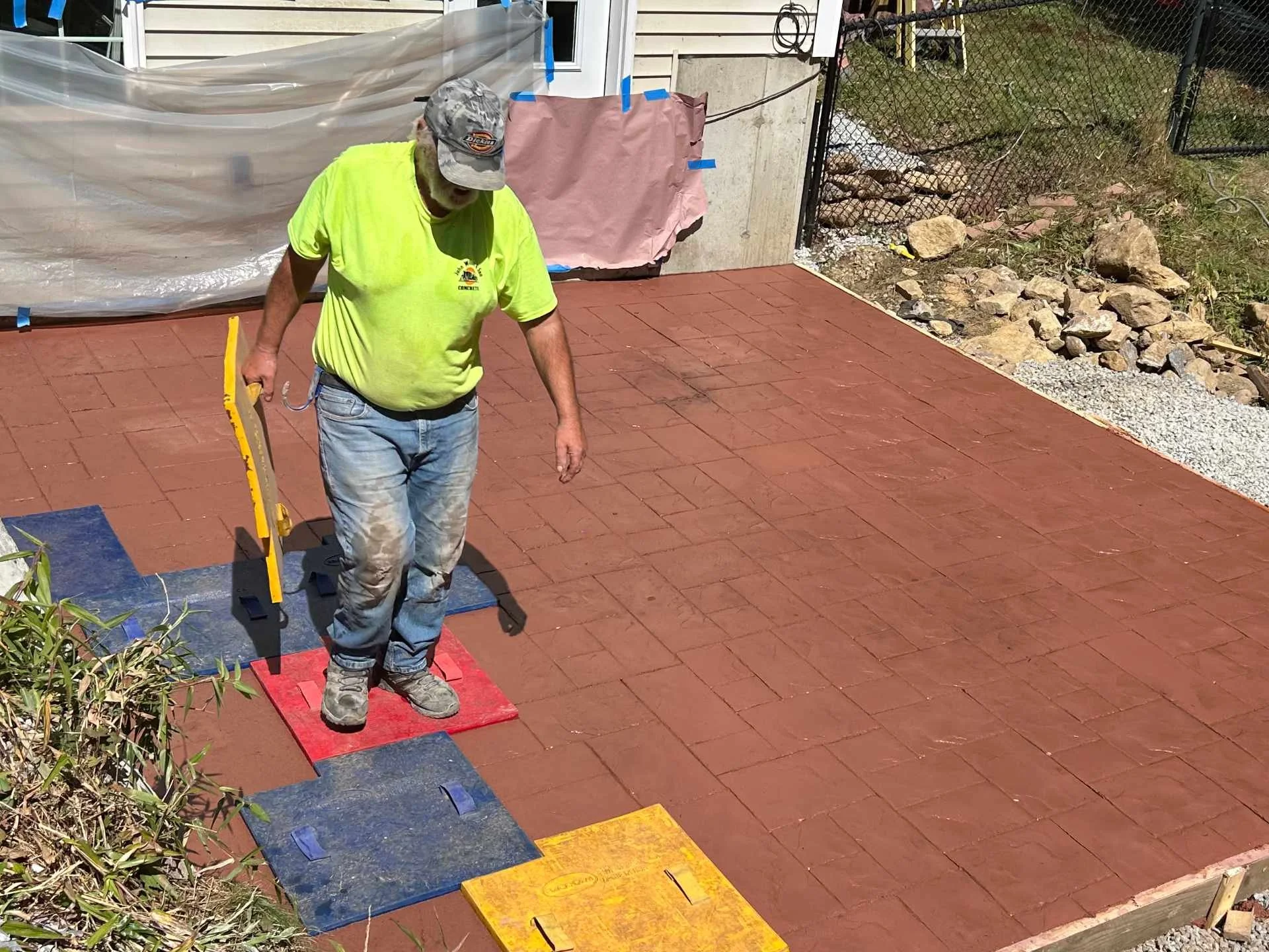 A person wearing a yellow shirt, jeans, and a cap is standing on a concrete stamp installing a decorative stamped concrete slab. 