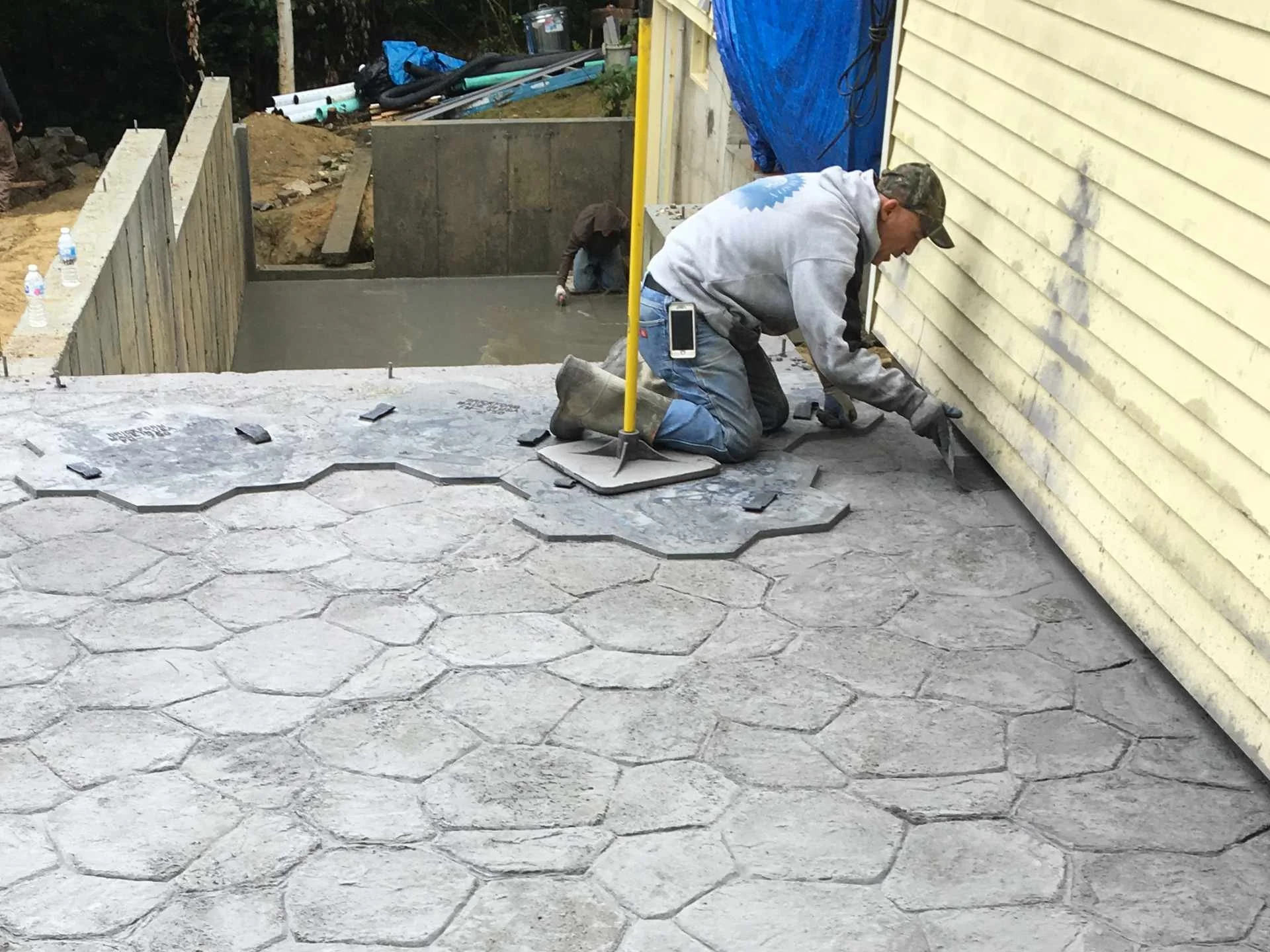 Two construction workers are pouring and leveling concrete for a new outdoor area, with one kneeling and stamping the wet concrete with a decorative pattern and the other working near the house siding.