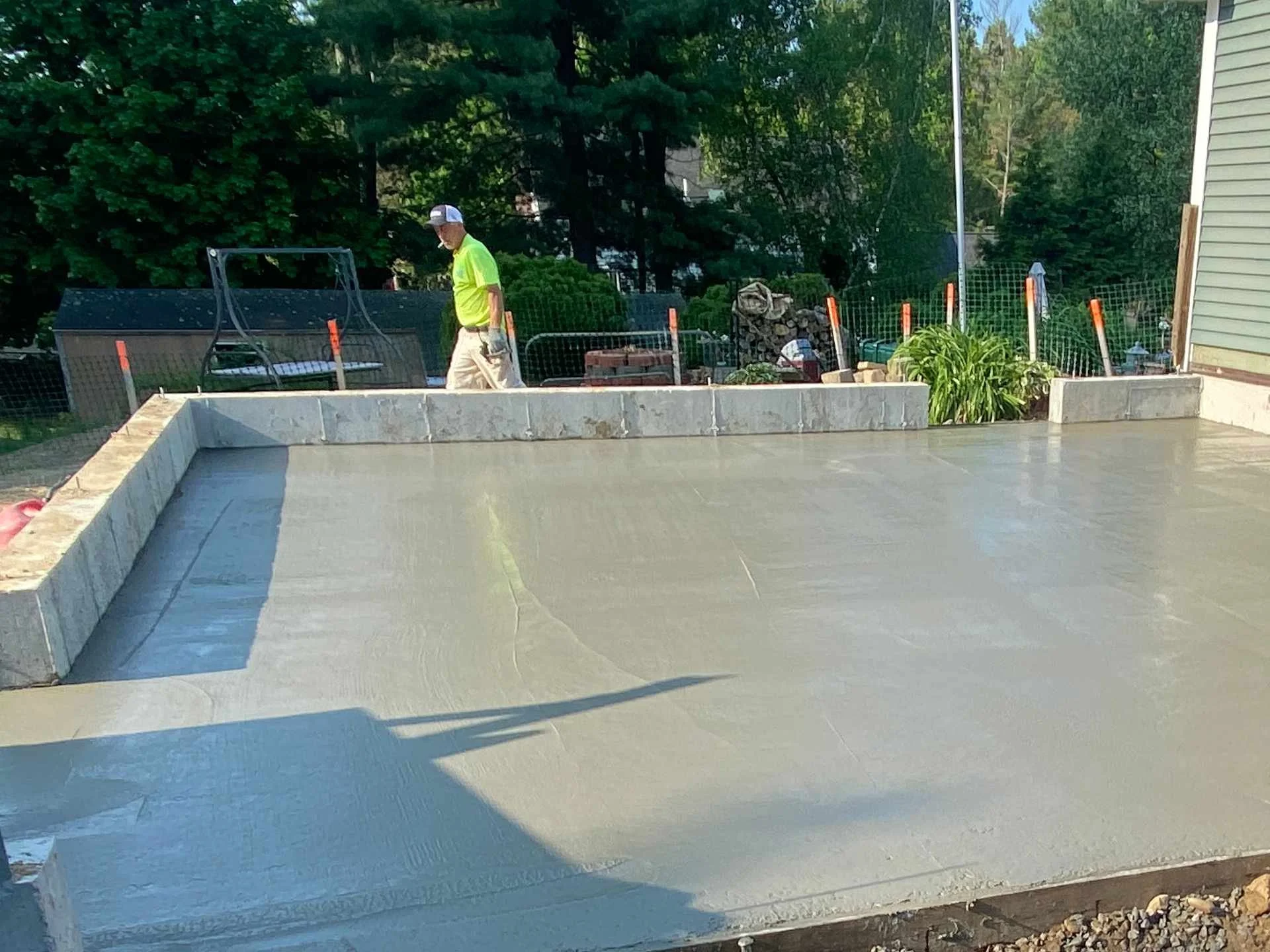 A man in a neon yellow shirt and khaki pants is walking on a freshly poured concrete surface in a backyard. He is wearing a baseball cap and gloves, with trees and a house in the background.