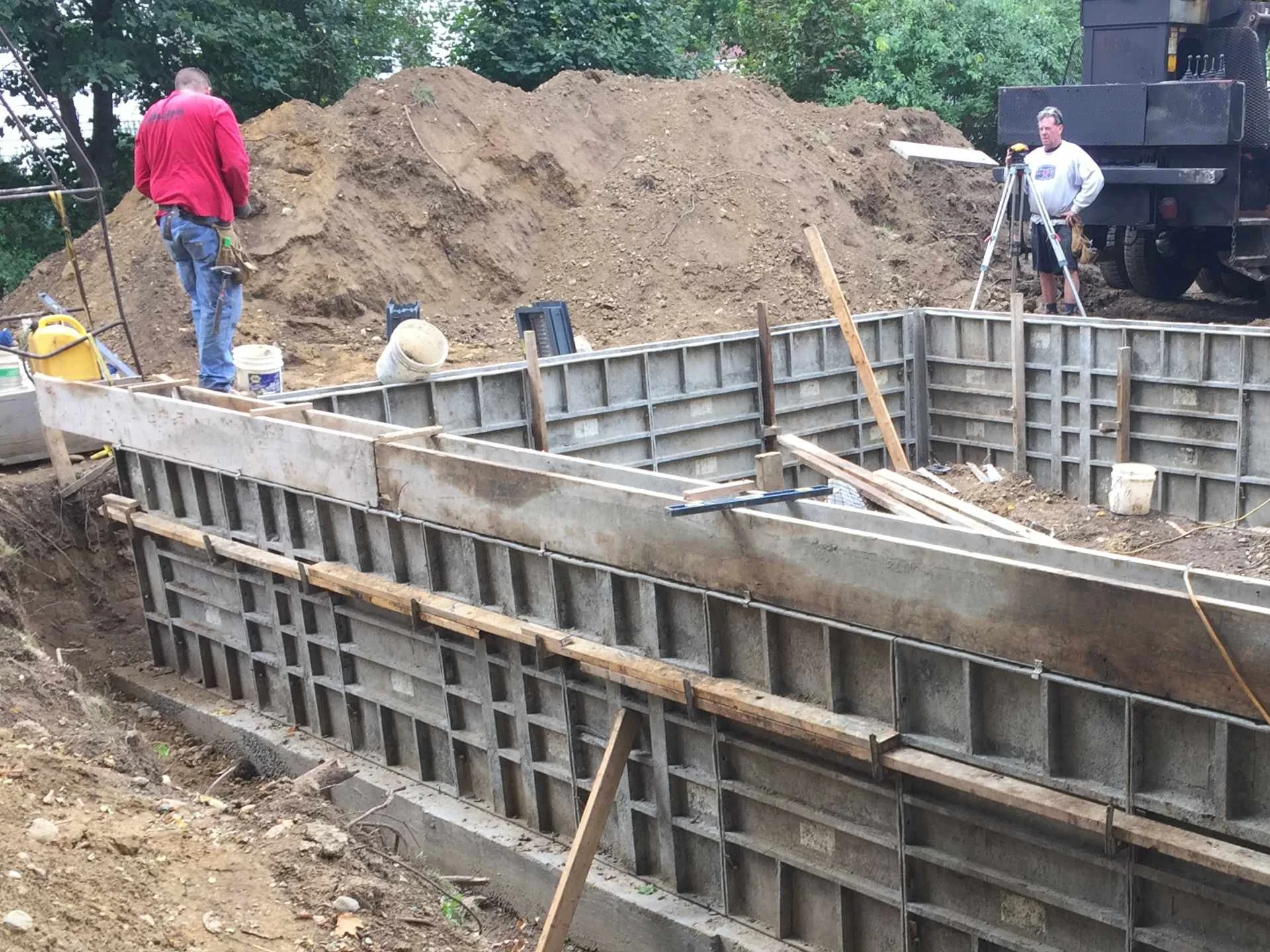 Construction site with two workers, one in a red jacket and blue jeans, and the other in a white sweatshirt, operating surveying equipment near a deep excavation with concrete formwork. There is a large dirt mound behind them and construction tools s