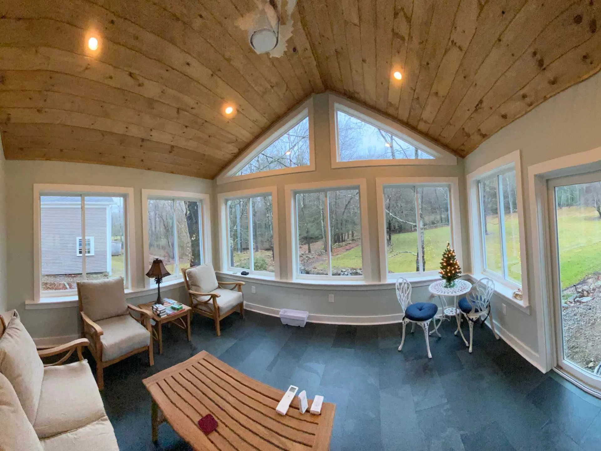 Living room with large window wall and a wooden ceiling with recessed lighting, decorated for Christmas with a small decorated tree, beige armchairs, a dining table with white chairs, and a view of a backyard with trees.