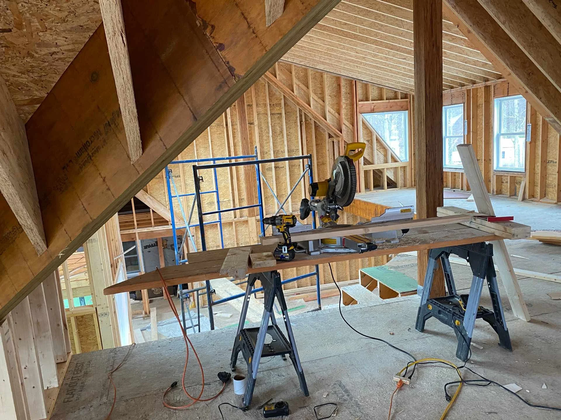 Interior of a house under construction, showing exposed wooden framing, windows, and construction tools including saws and ladders.