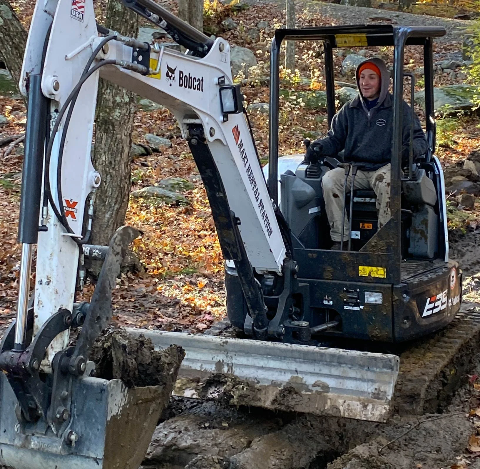 A person operating a Bobcat excavator in an outdoor wooded area with fallen leaves and trees.