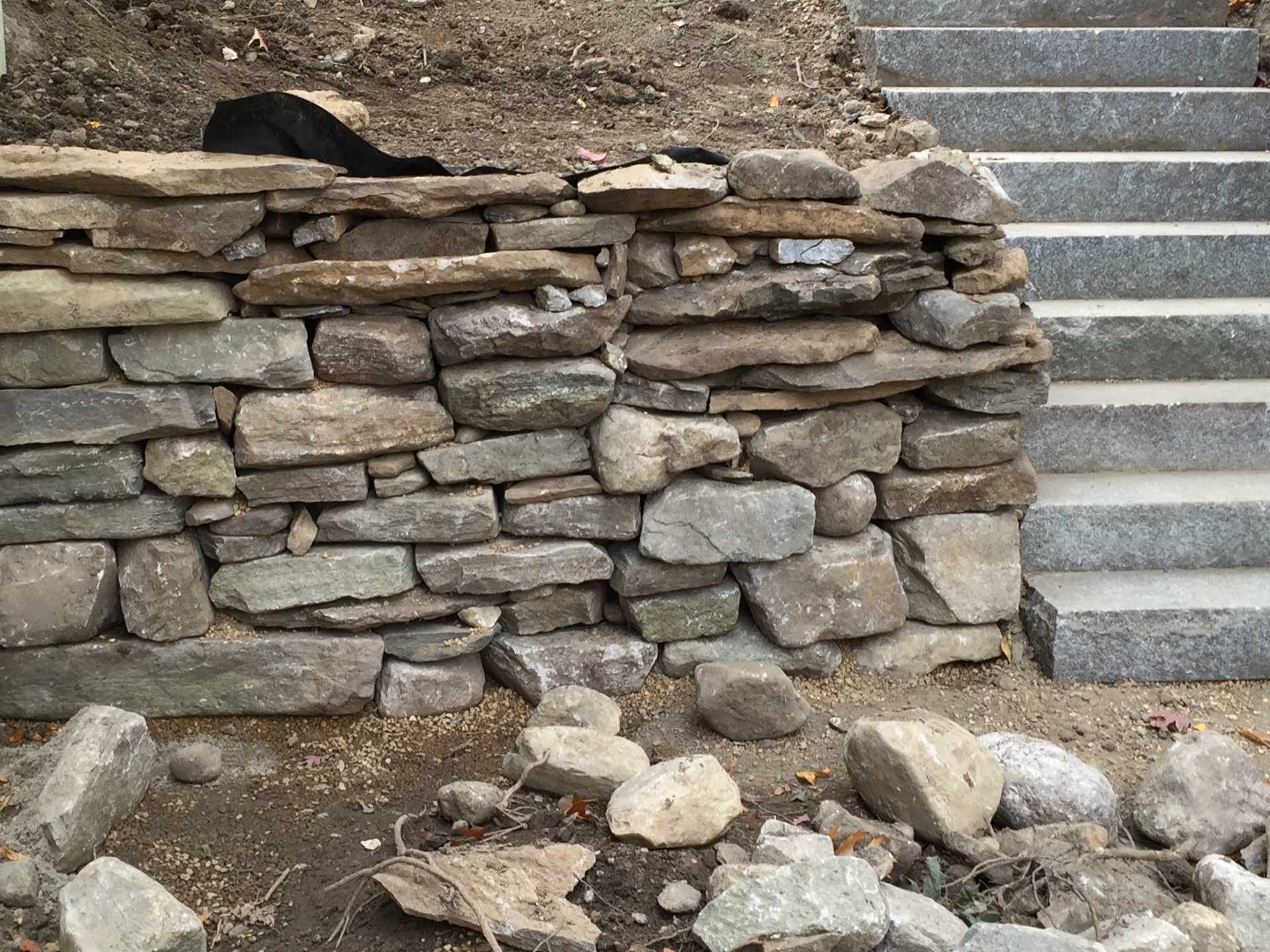 A stone retaining wall with a set of granite steps to the right and loose rocks and dirt in front of it.