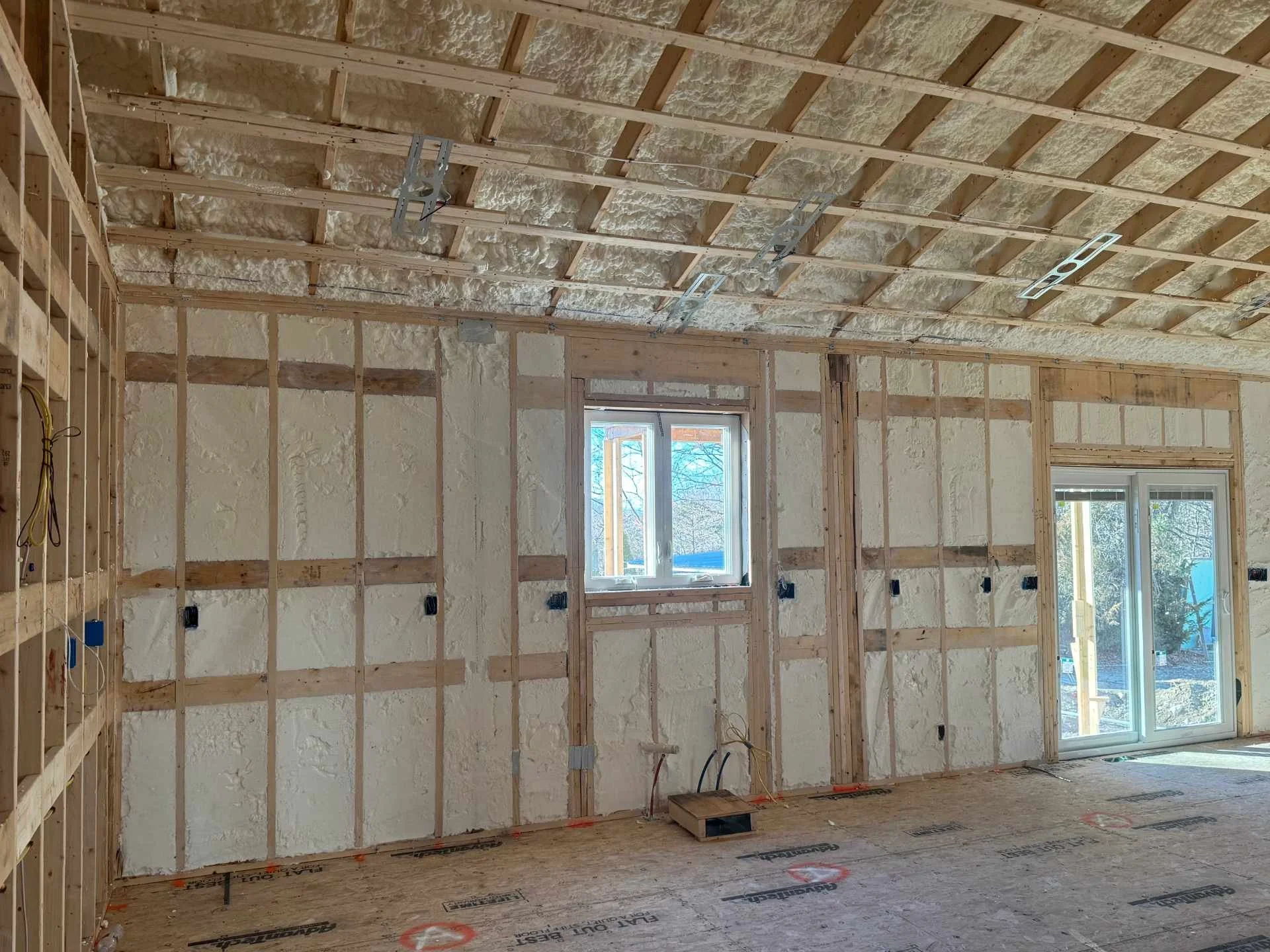 Interior of a house under construction with exposed wooden framing, closed-cell spray foam insulation on the ceiling and walls, electrical wiring, and windows for natural light.