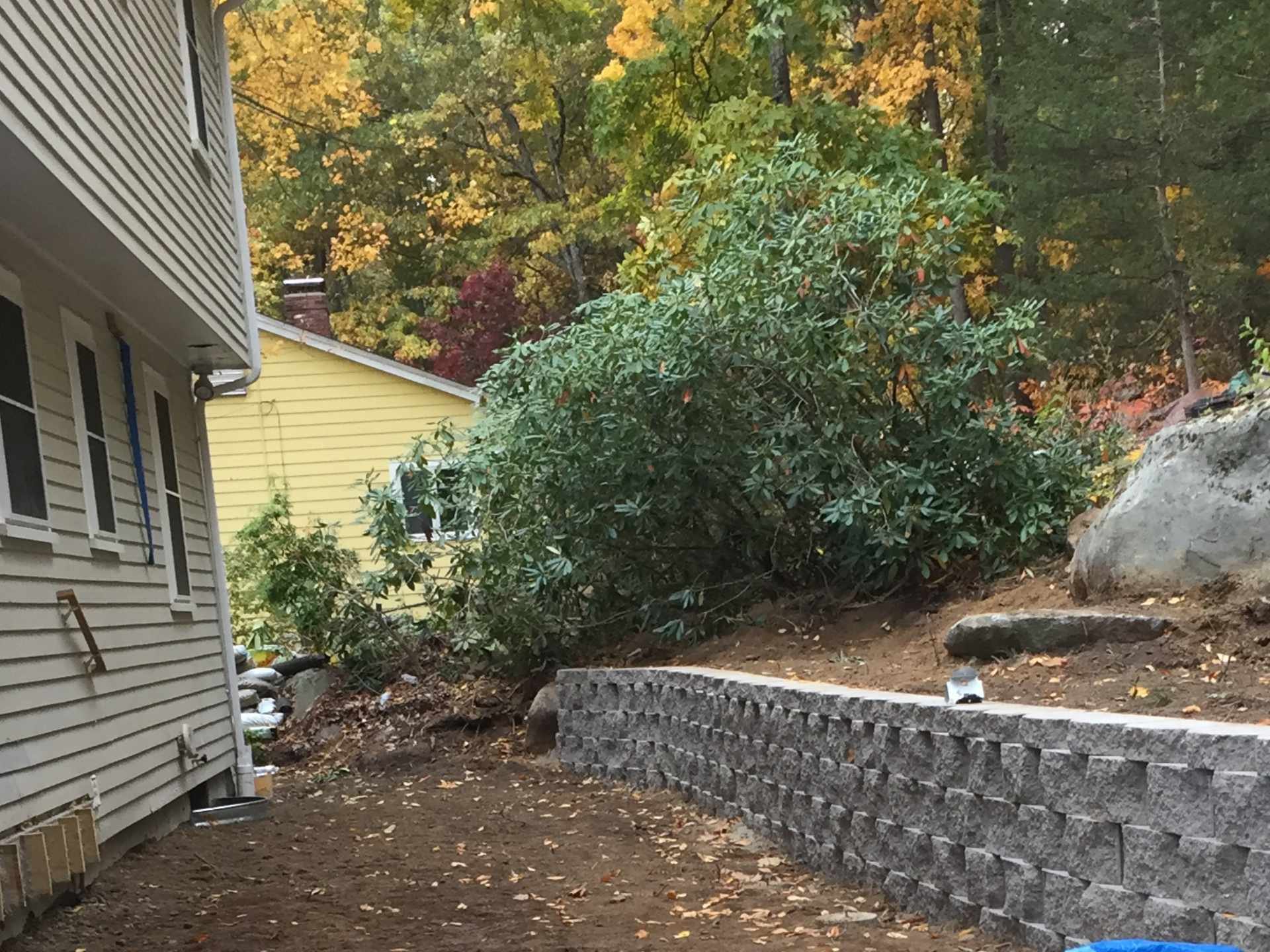 A backyard area with a stone retaining wall and dirt ground, adjacent to a house with beige siding and a window, surrounded by trees with autumn-colored leaves.
