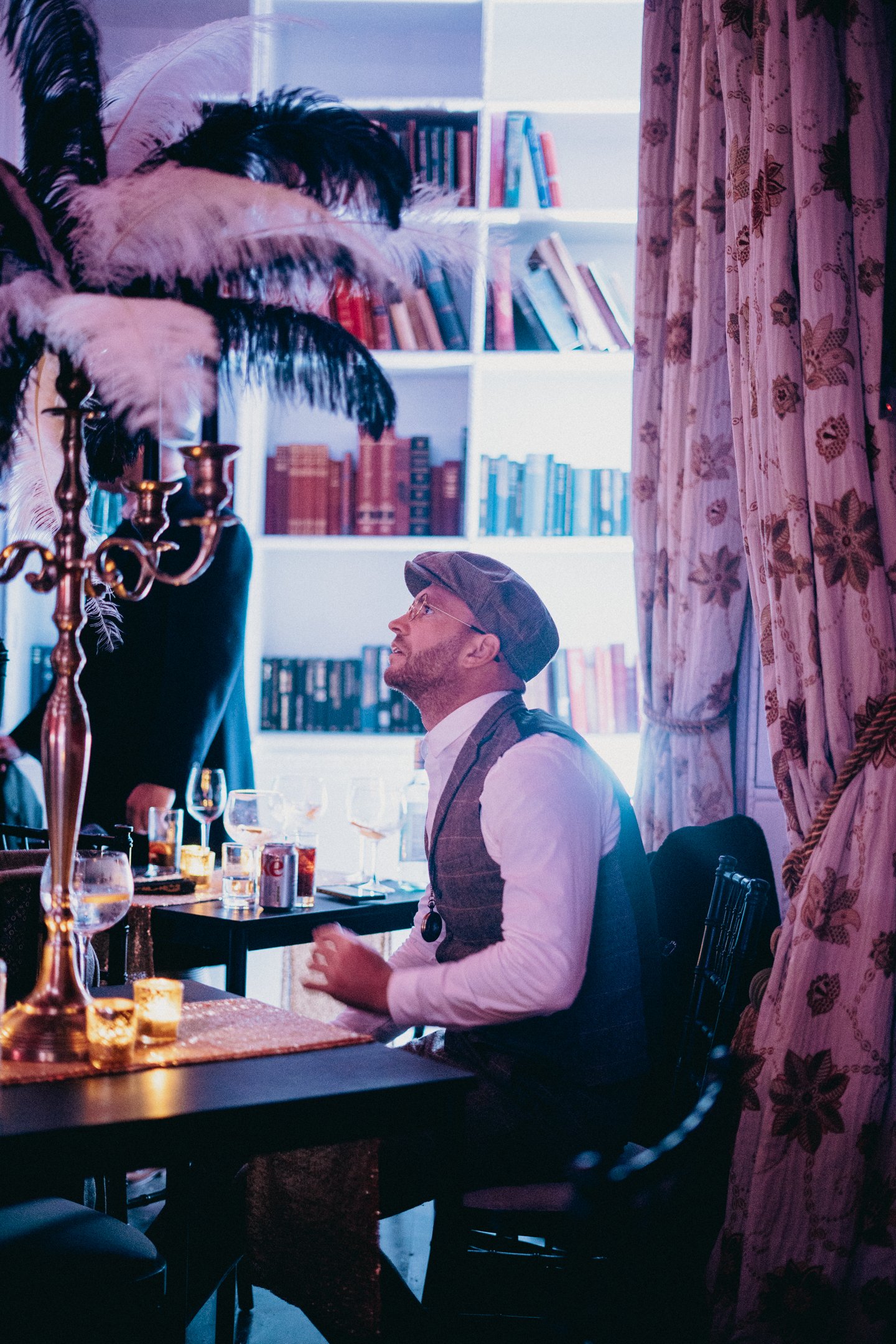 A man with glasses, wearing a vest and a brown hat, sitting at a table in a cozy, well-lit room with shelves of books and floral curtains. The table has glasses, a soda can, and candle holders. A woman with dark clothing and feathers in her hair is standing nearby.