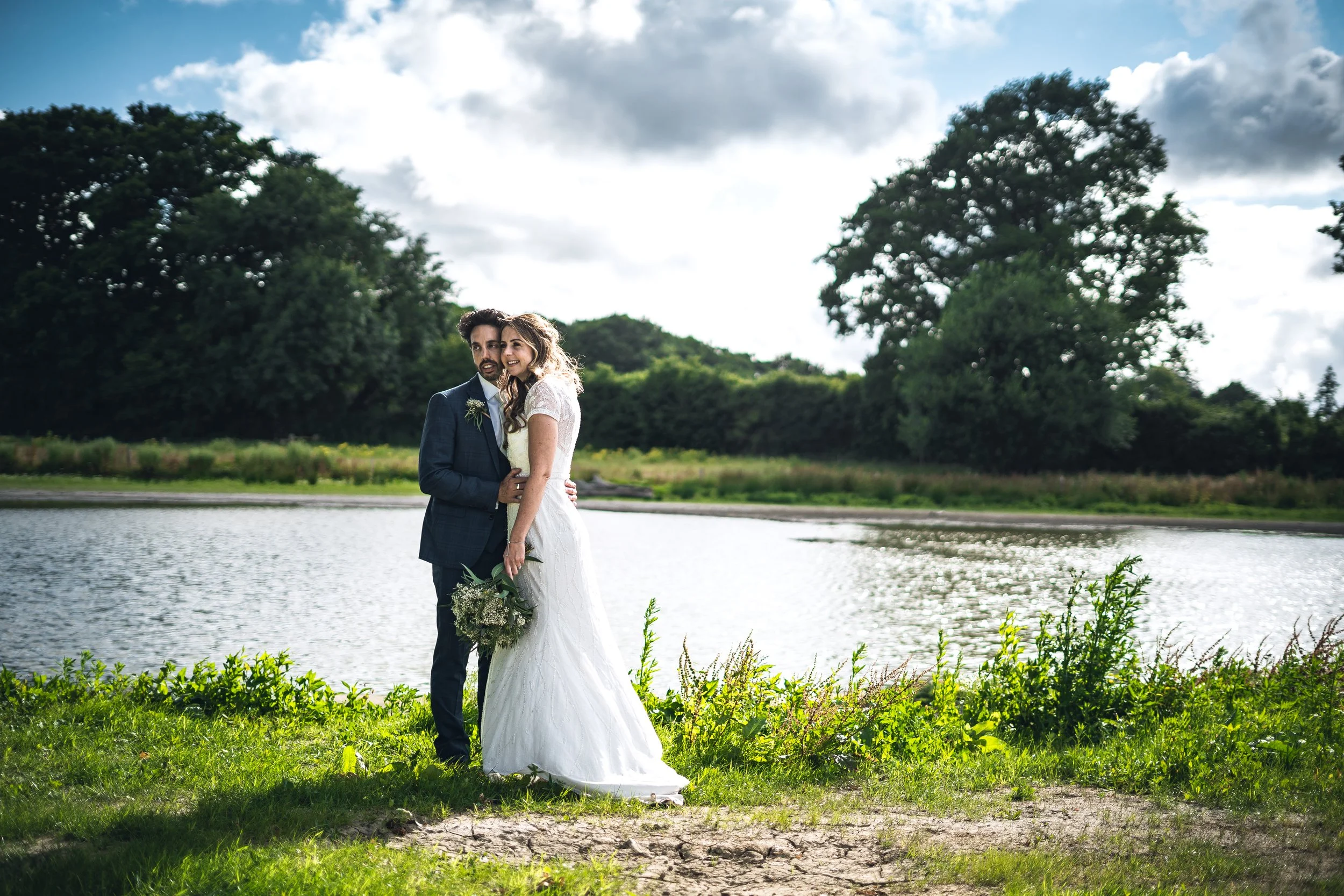 A bride and groom standing together by a lake in a park, with trees and clouds in the background, during a wedding photoshoot.