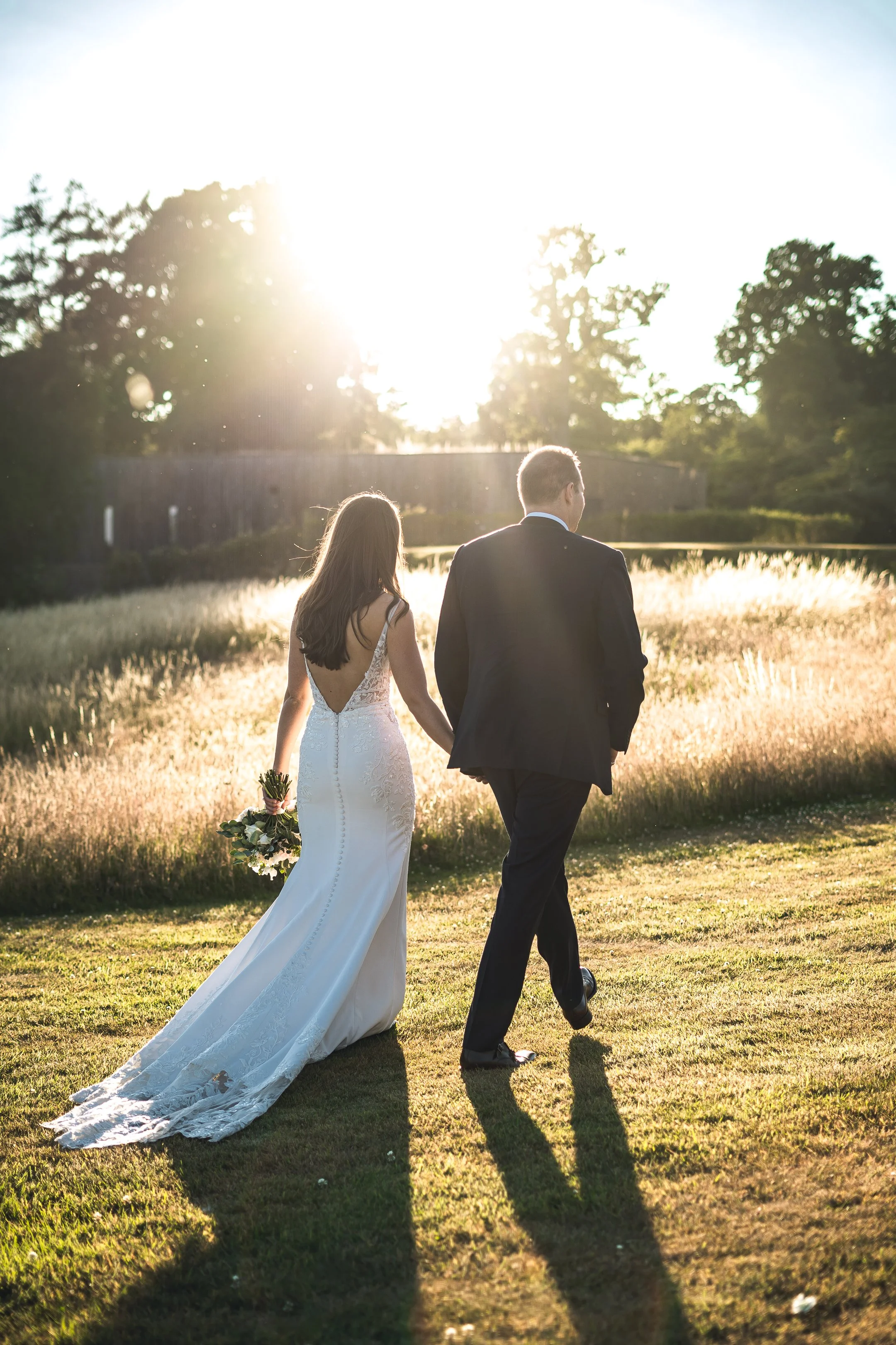 A bride and groom walking outdoors on a sunny day, holding hands, with the bride holding a bouquet of flowers. The bride is in a white gown and the groom in a black suit.