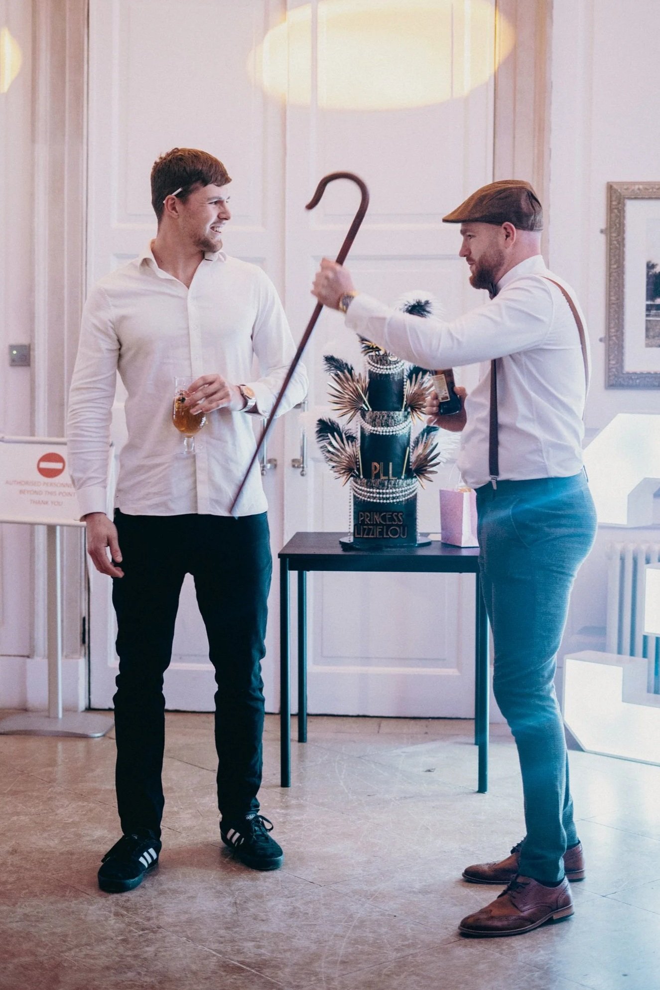 Two men stand and smile at each other during a celebration, with a decorated cake on a table between them. The man on the left wears a white shirt, black pants, and is holding a drink. The man on the right wears a white shirt, gray pants, a flat cap, and suspenders. The cake is black and gold with a crown decoration and the words "Princess Lizelou" written on it. The background features a framed picture and a transparent sign with a red no-entry symbol.