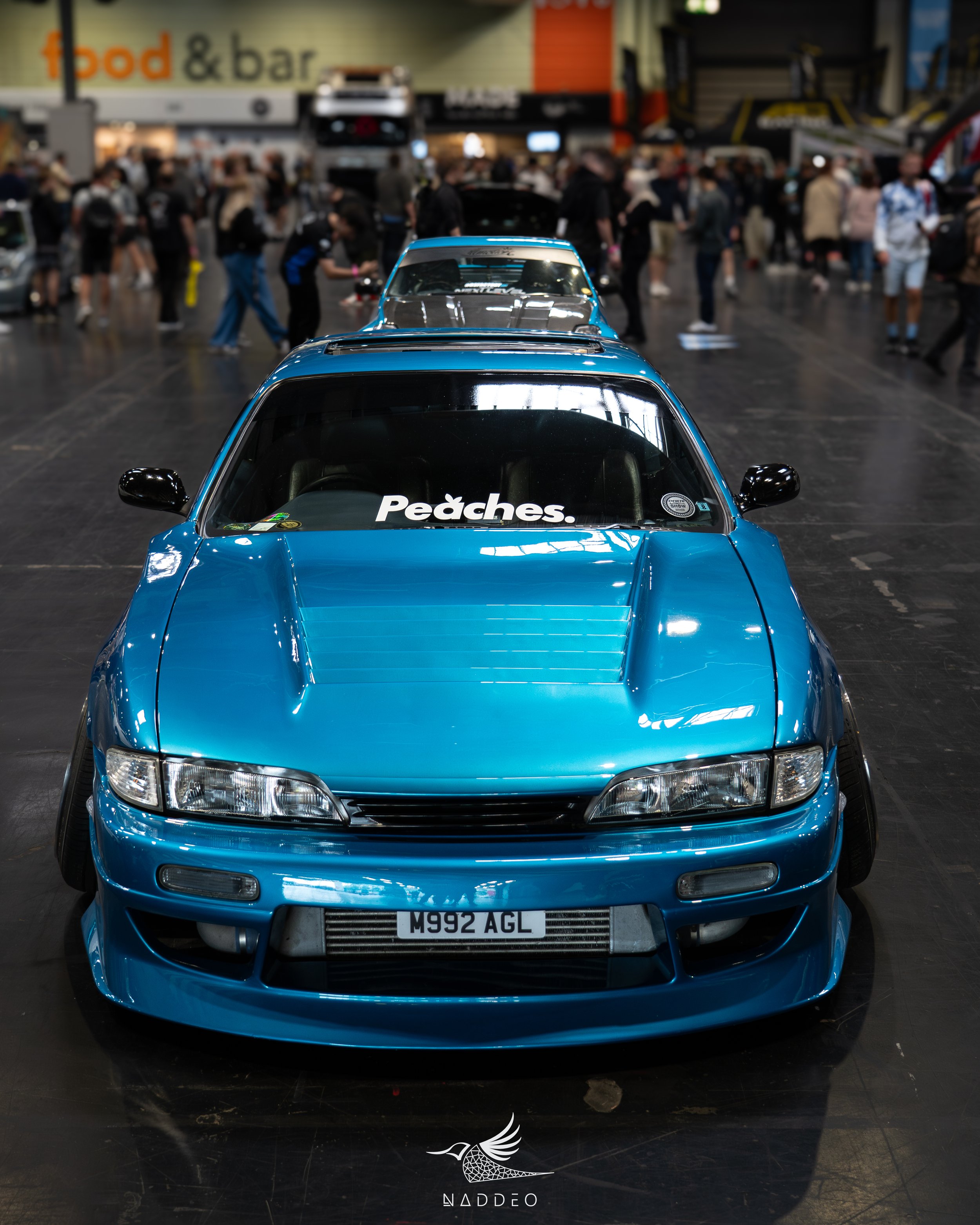 Front view of a blue sports car with a white sticker that says 'Pëdches' on the windshield, displayed at an indoor car exhibition with people walking around in the background.