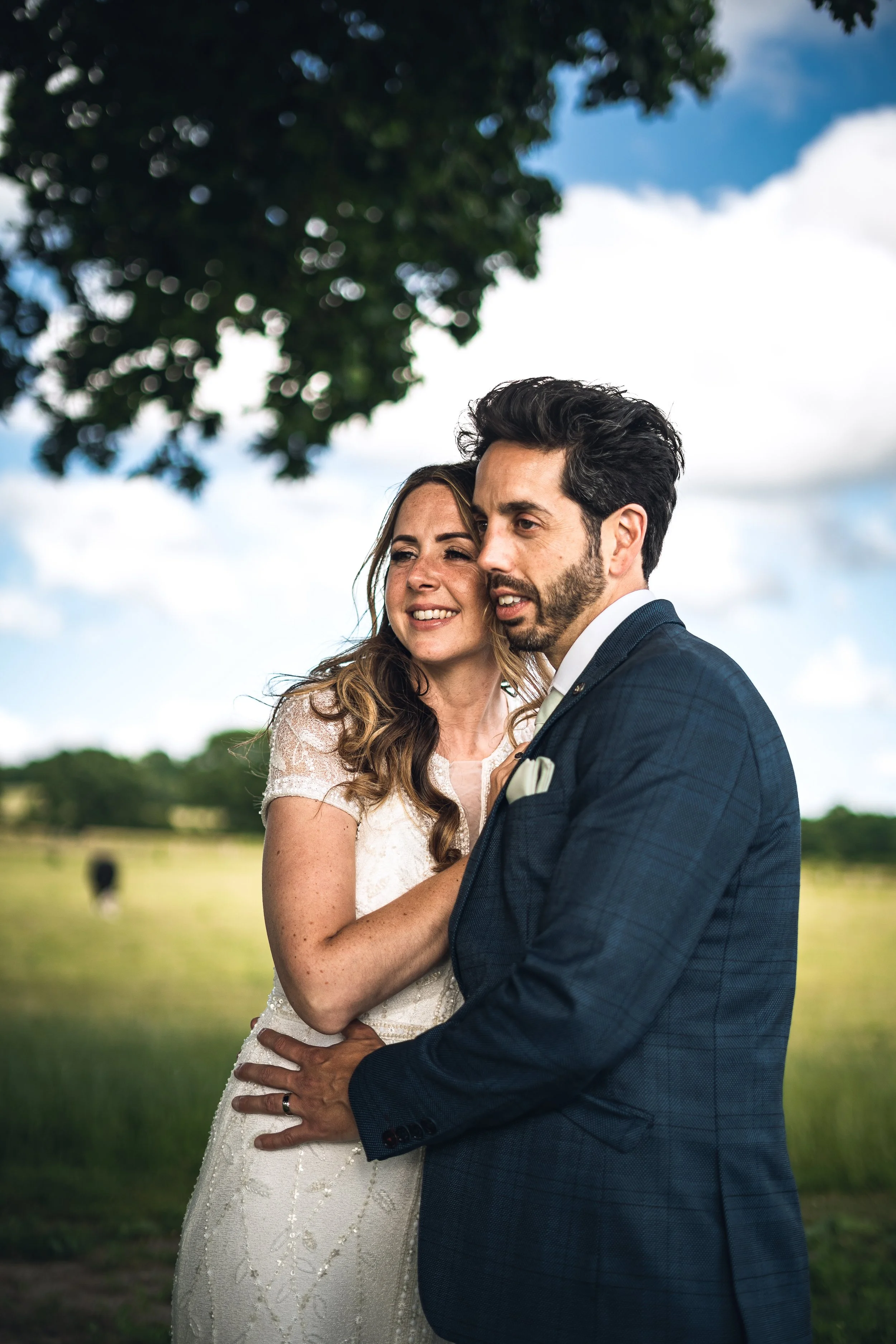 A couple standing closely outdoors, loving embrace, smiling, with a tree and blue sky with white clouds in the background.