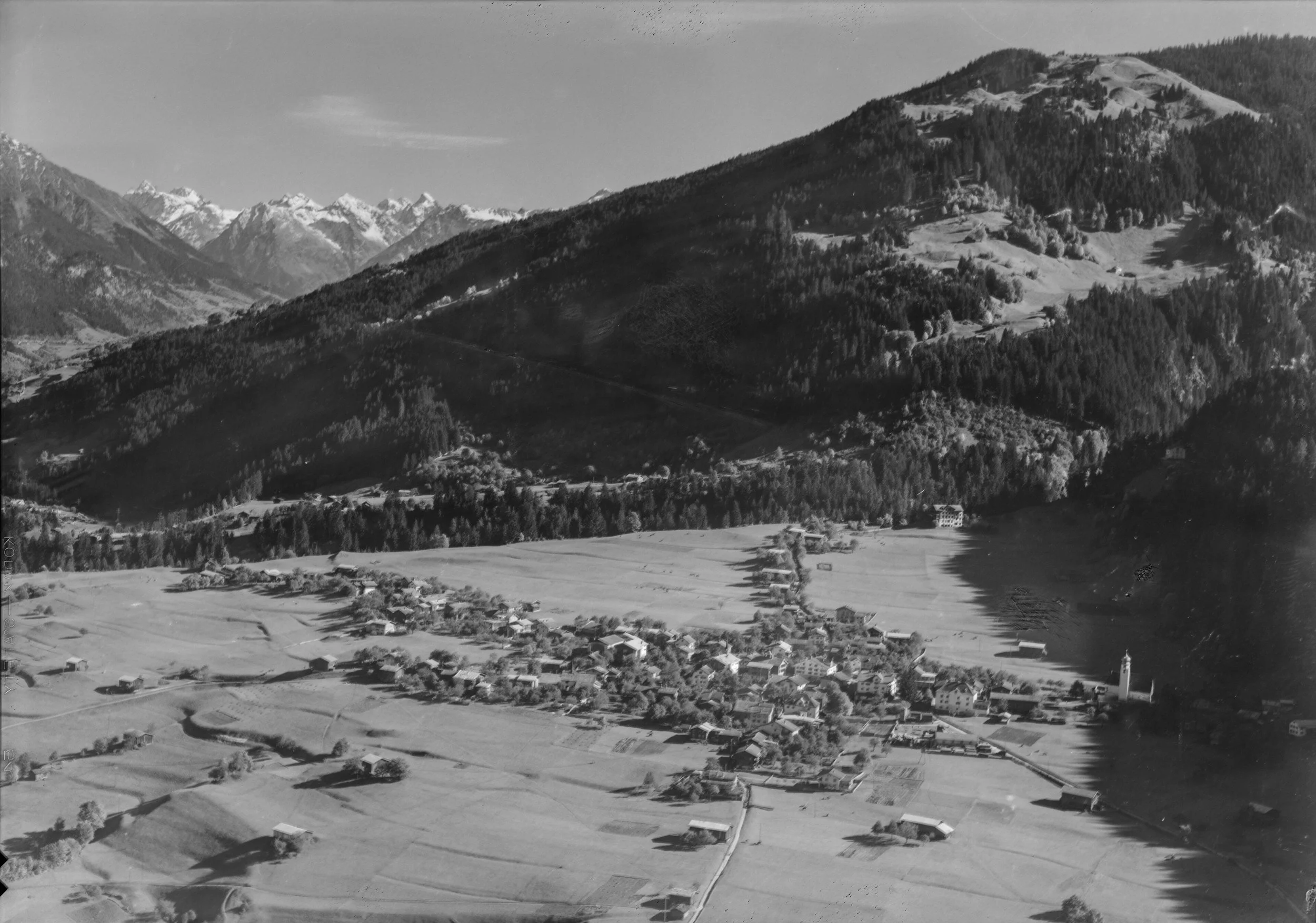 Schwarz-Weiß-Fotografie einer Berglandschaft mit kleinen Dörfern im Tal, umgeben von Wäldern und hohen Bergen im Hintergrund.