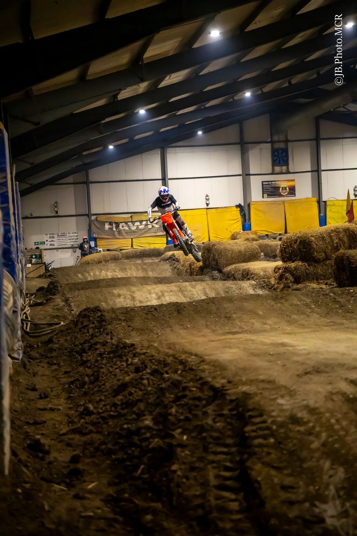 An indoor dirt bike or motocross track with a rider in mid-air, wearing a helmet and riding gear, surrounded by hay bales for safety, under a roof with spotlights.