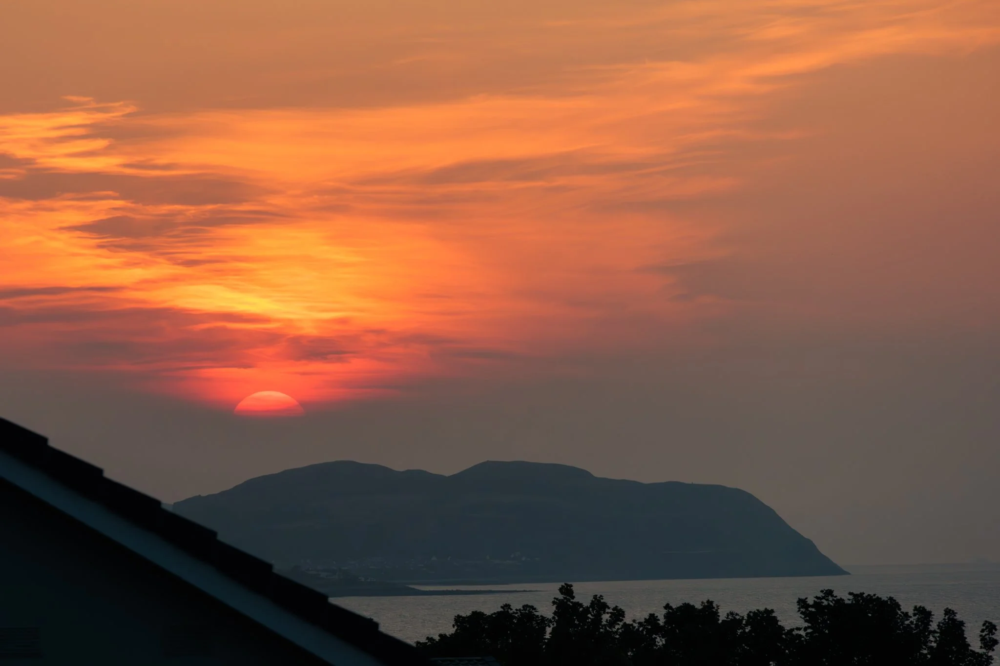 Sun setting behind mountains with a colorful sky of orange, pink, and purple hues, viewed from a vantage point that includes a silhouette of a building roof and trees in the foreground.