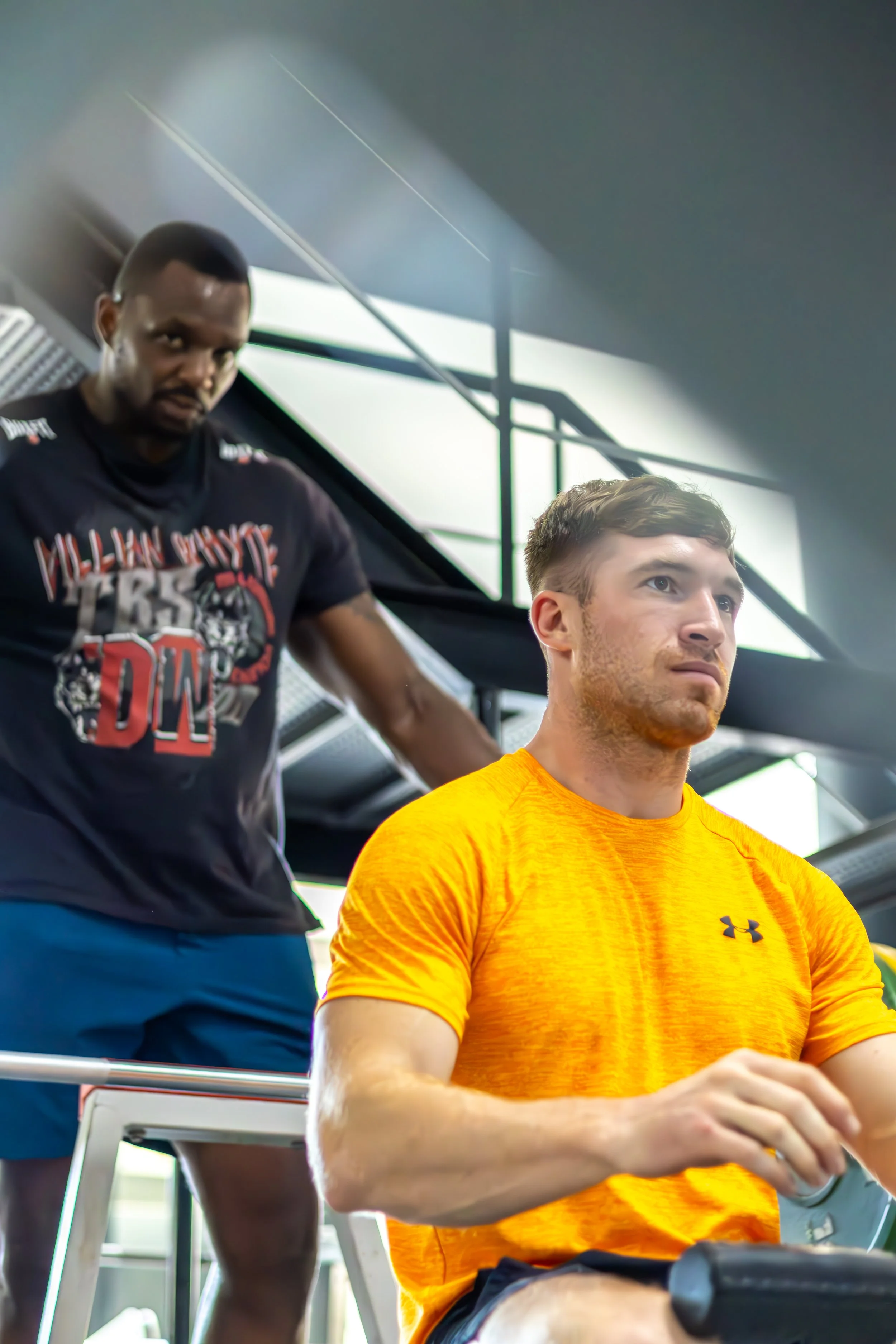 A man with a beard and short hair wearing a bright yellow athletic shirt sitting on a rowing machine in a gym, with another man in the background working out.