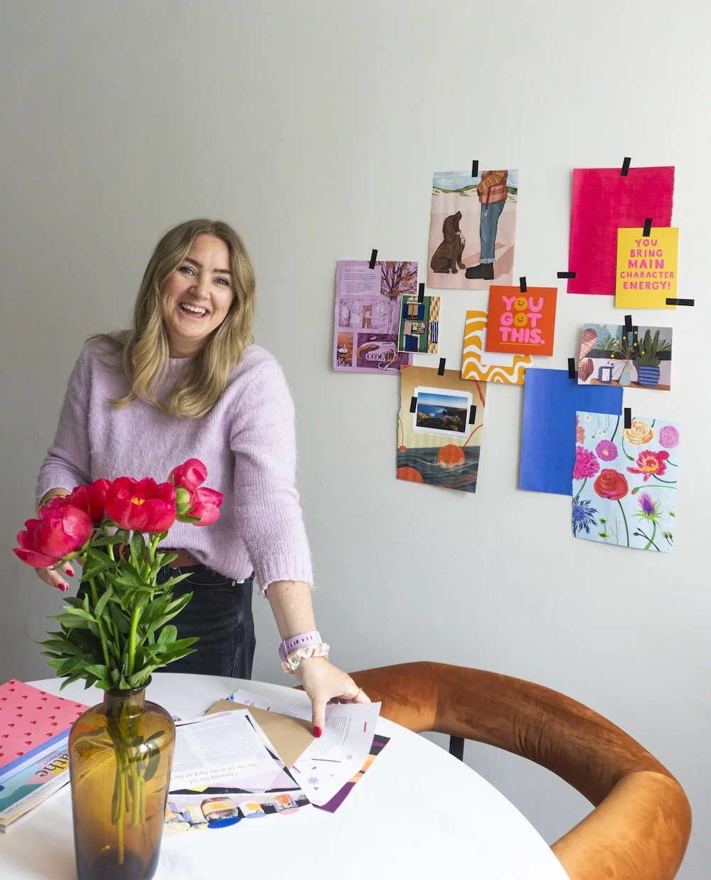 A smiling woman in a pink sweater standing at a table with pink flowers in a brown vase and papers, in a room with a wall decorated with colorful postcards and illustrations taped with black tape.