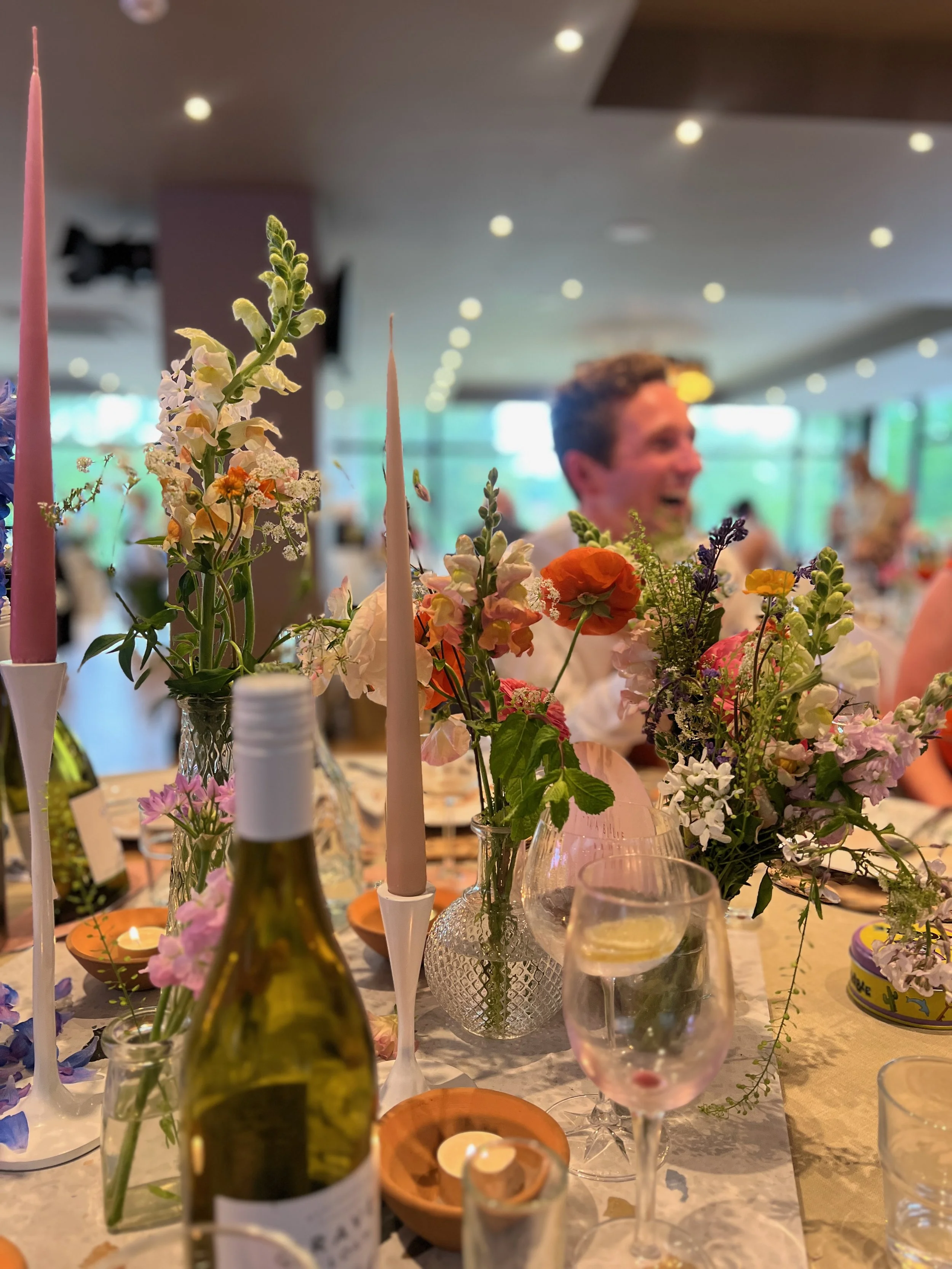 A table decorated with colorful flowers, candles, and glassware at a celebration or gathering, with a smiling man in the background.