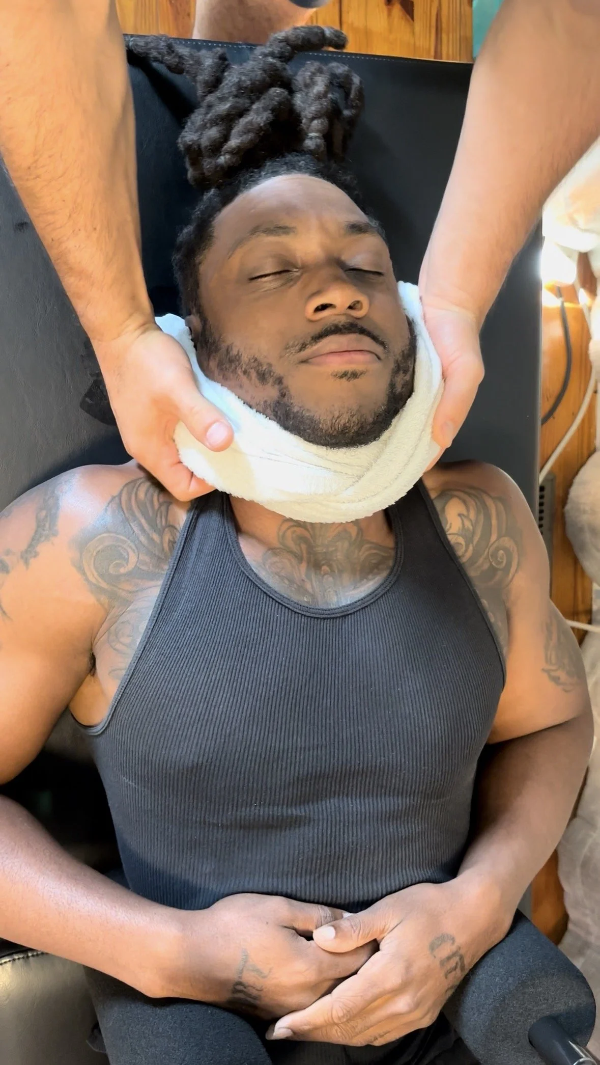 A man with closed eyes receiving a neck massage from a person with hands on his neck, lying on a massage table with a towel underneath his head, wearing a black sleeveless shirt, with tattoos on his arms and chest.
