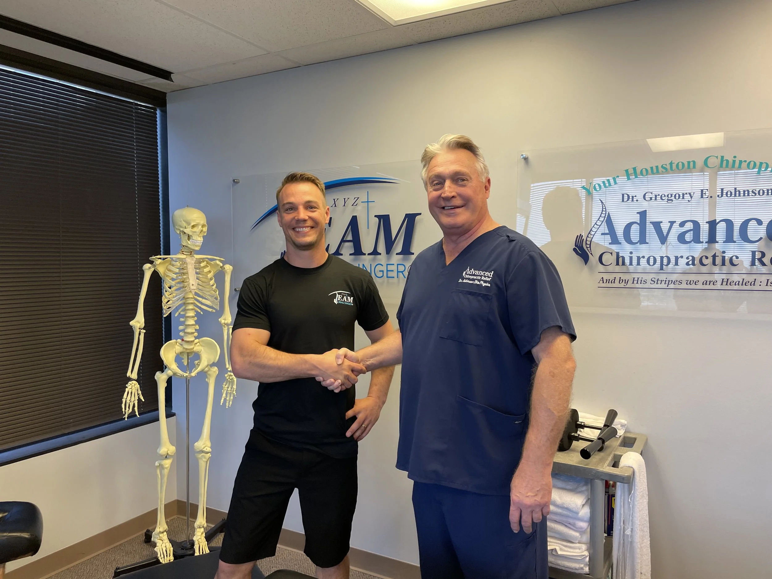 Two men shaking hands in a chiropractic office, with a human skeleton model beside them and a sign behind them that reads "Your Houston Chiropractor Dr. Gregory L. Johnson".