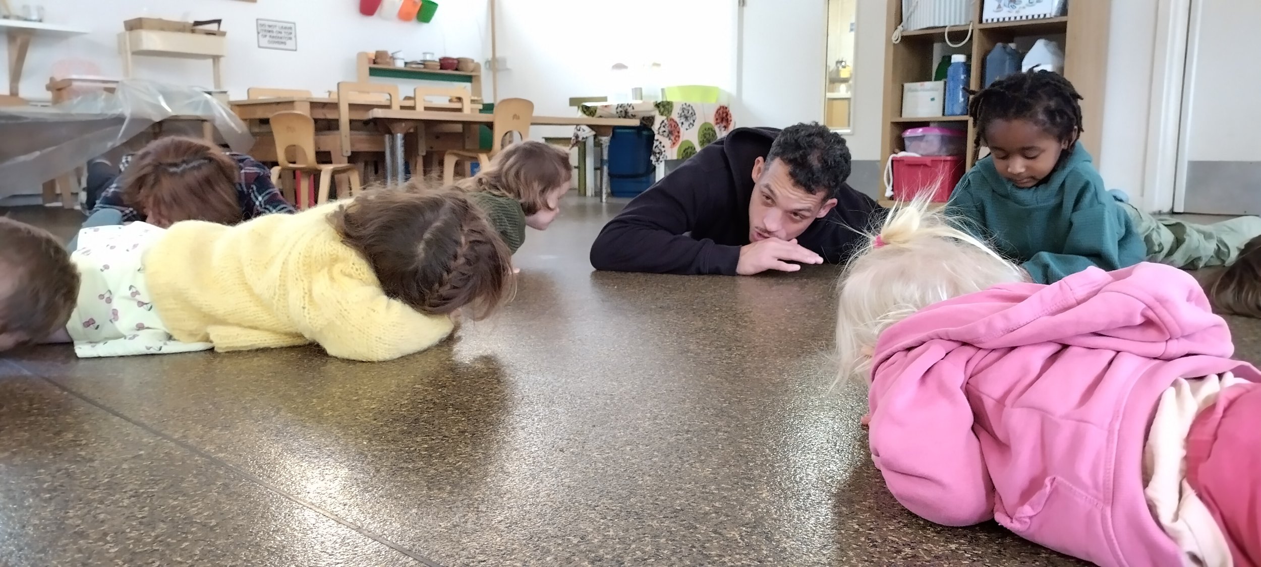 A group of children lying on the floor with their heads down, listening to an adult male who is lying on his stomach, resting his head on his hand, in a classroom or activity room.