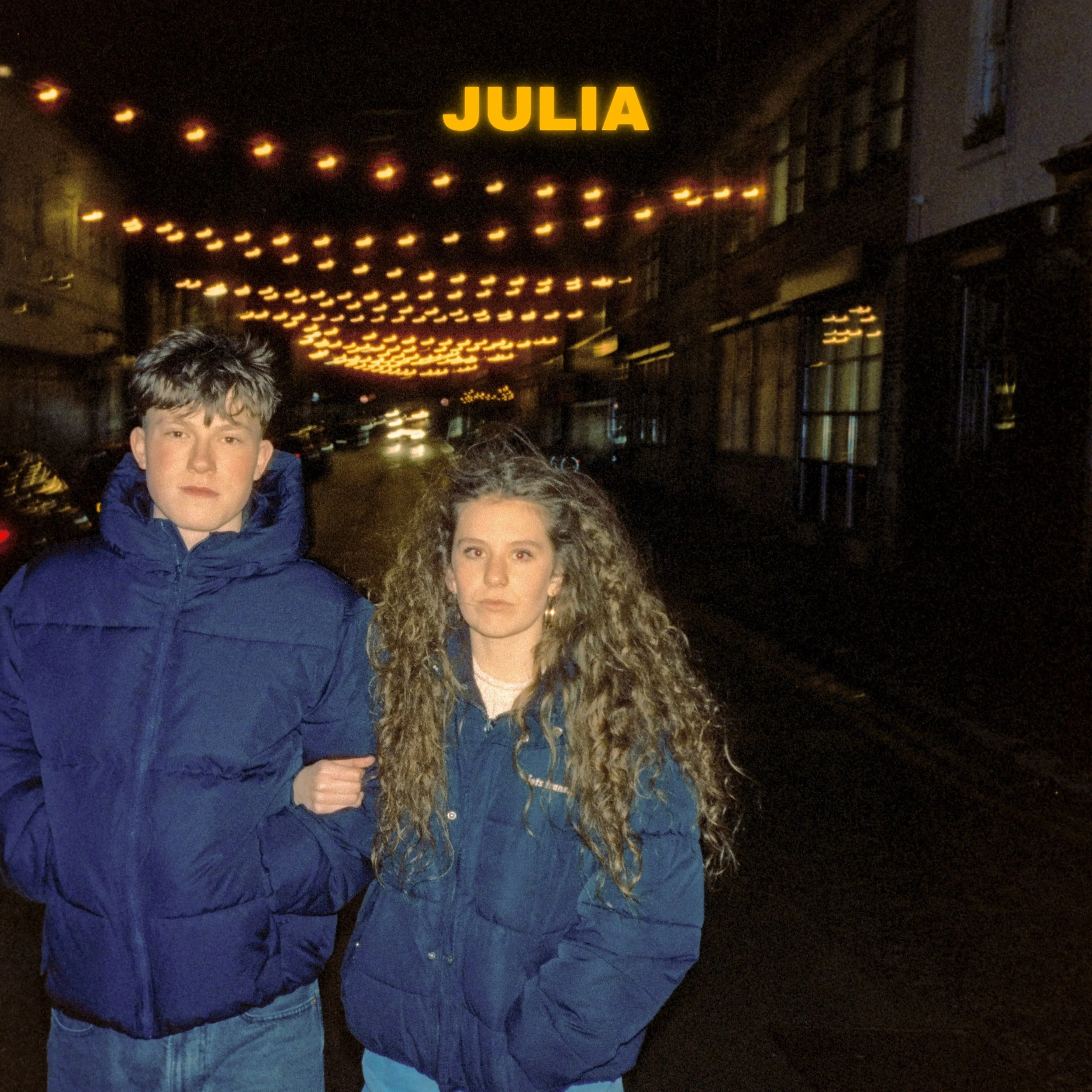 Nighttime street scene with string lights and a glowing yellow sign that says JULIA, three young people in winter jackets, partially visible faces, standing close together.