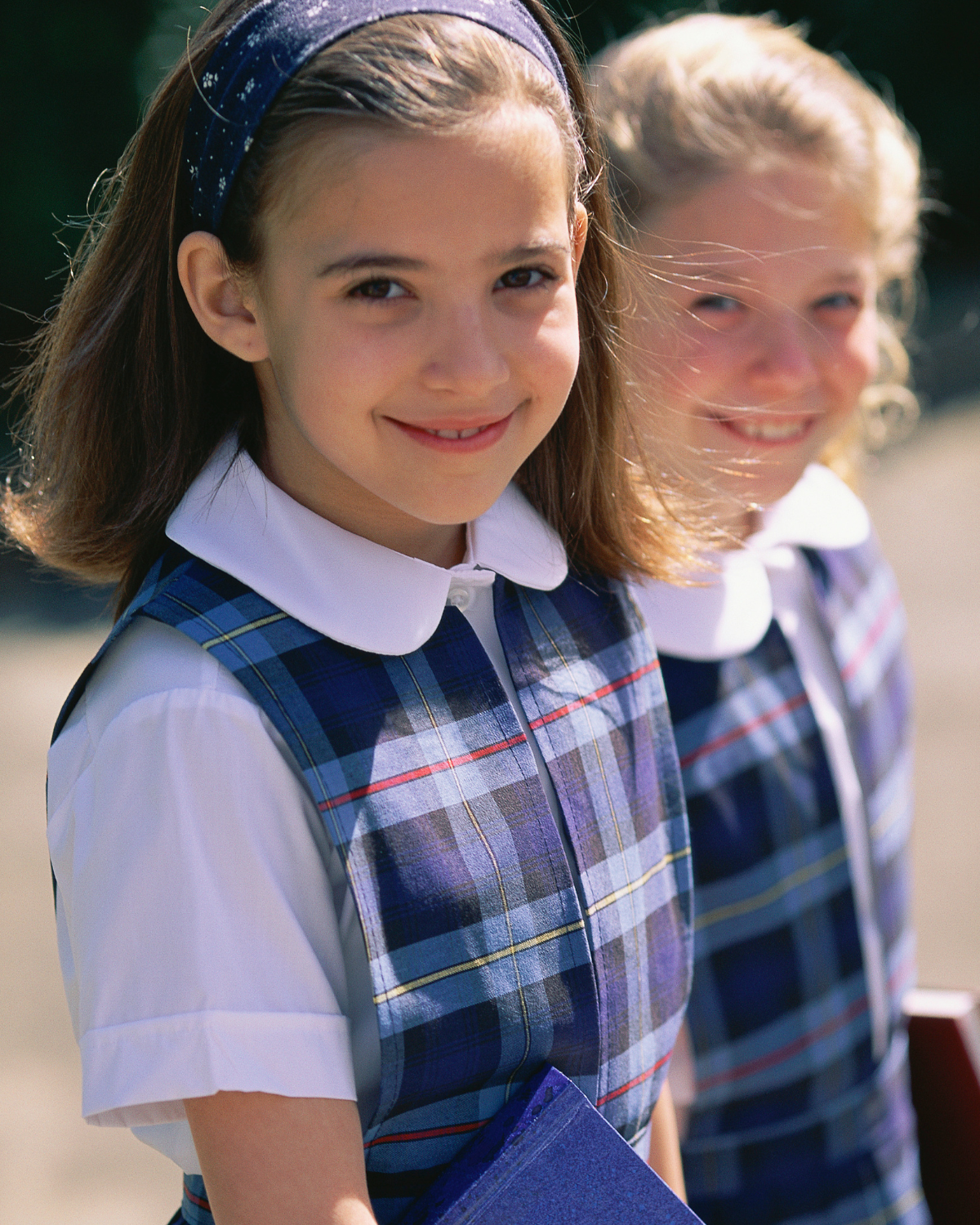 Two young girls wearing school uniforms with plaid vests and white collared shirts, smiling outdoors in sunlight.