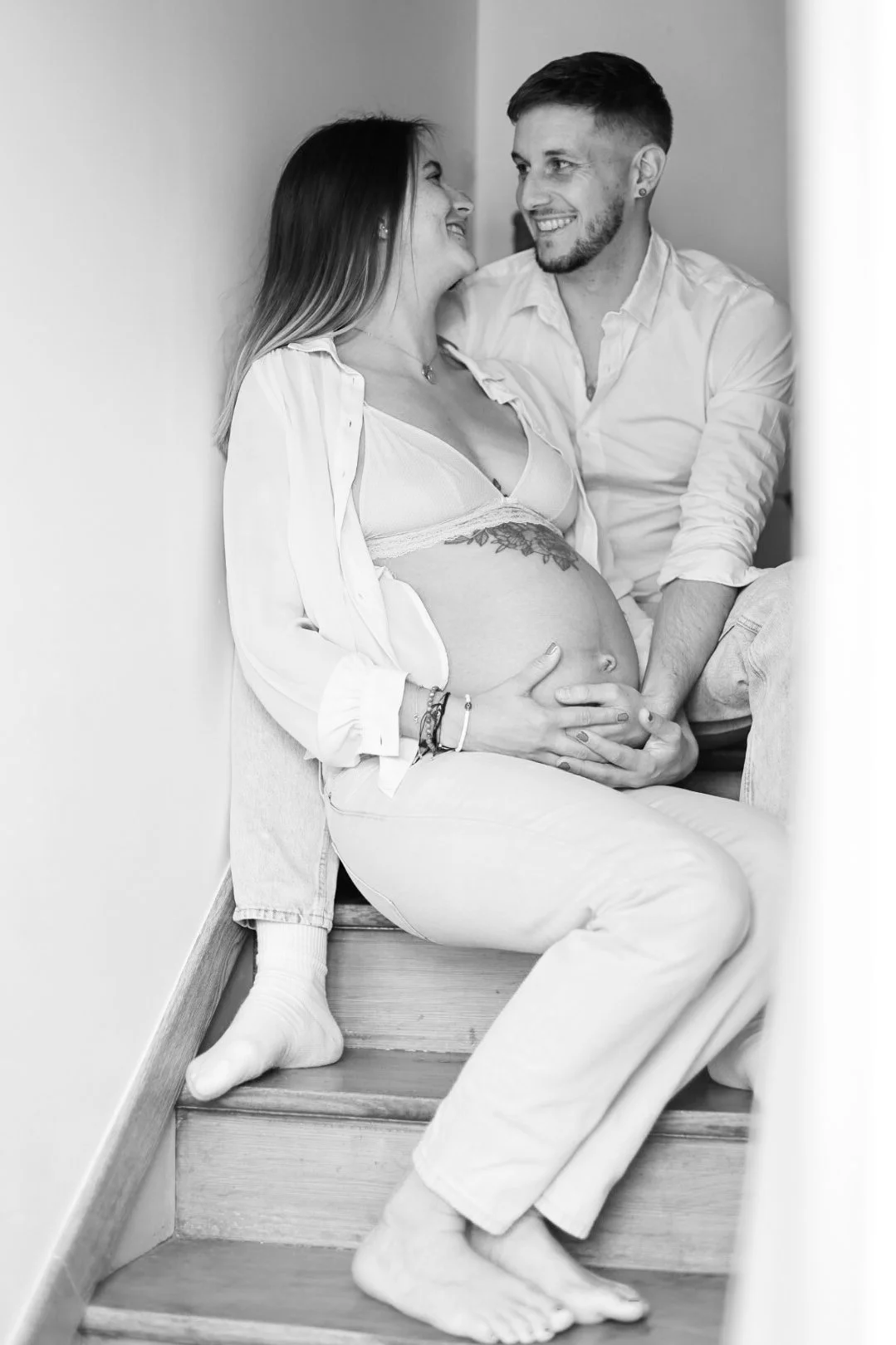 Une femme enceinte et un homme assis sur des escaliers, se regardant avec tendresse, en noir et blanc.