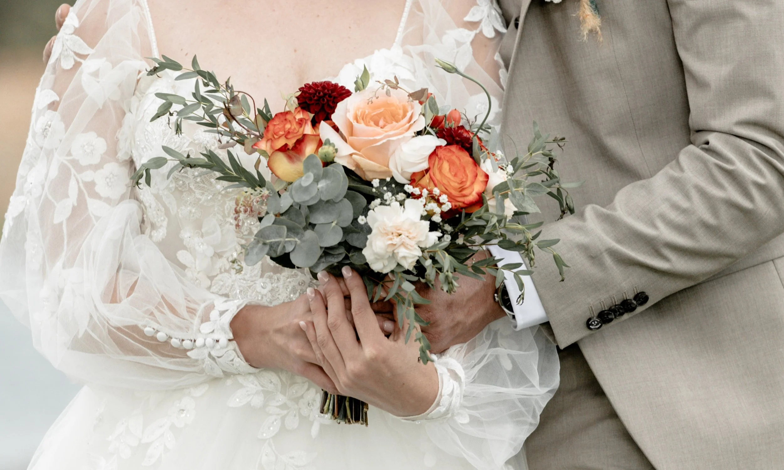 Un couple marié tenant un bouquet de fleurs, la femme en robe de mariage blanche en dentelle et le homme en costume gris avec poignets à boutons noirs.