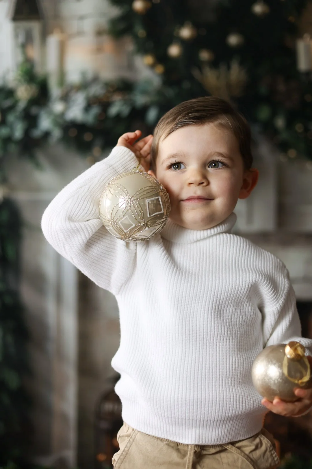 Un jeune garçon porte un pull blanc et tient deux décorations de Noël dorées, un dans chaque main, avec un sapin décoré en arrière-plan.