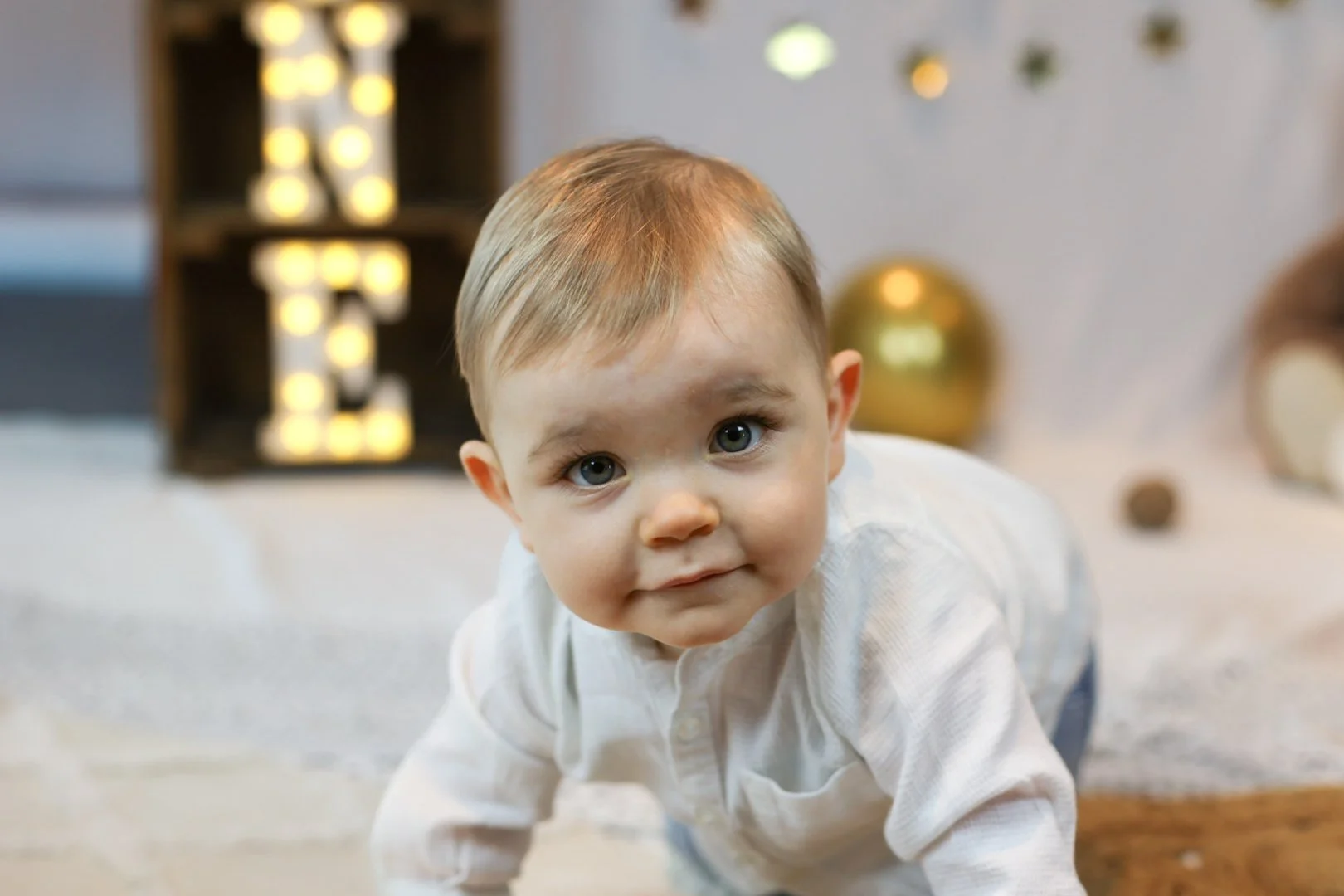 Un bébé qui rampe sur le sol, regardant vers l'avant, avec un décor de fête en arrière-plan, incluant des décorations dorées et une boîte avec des lumières.
