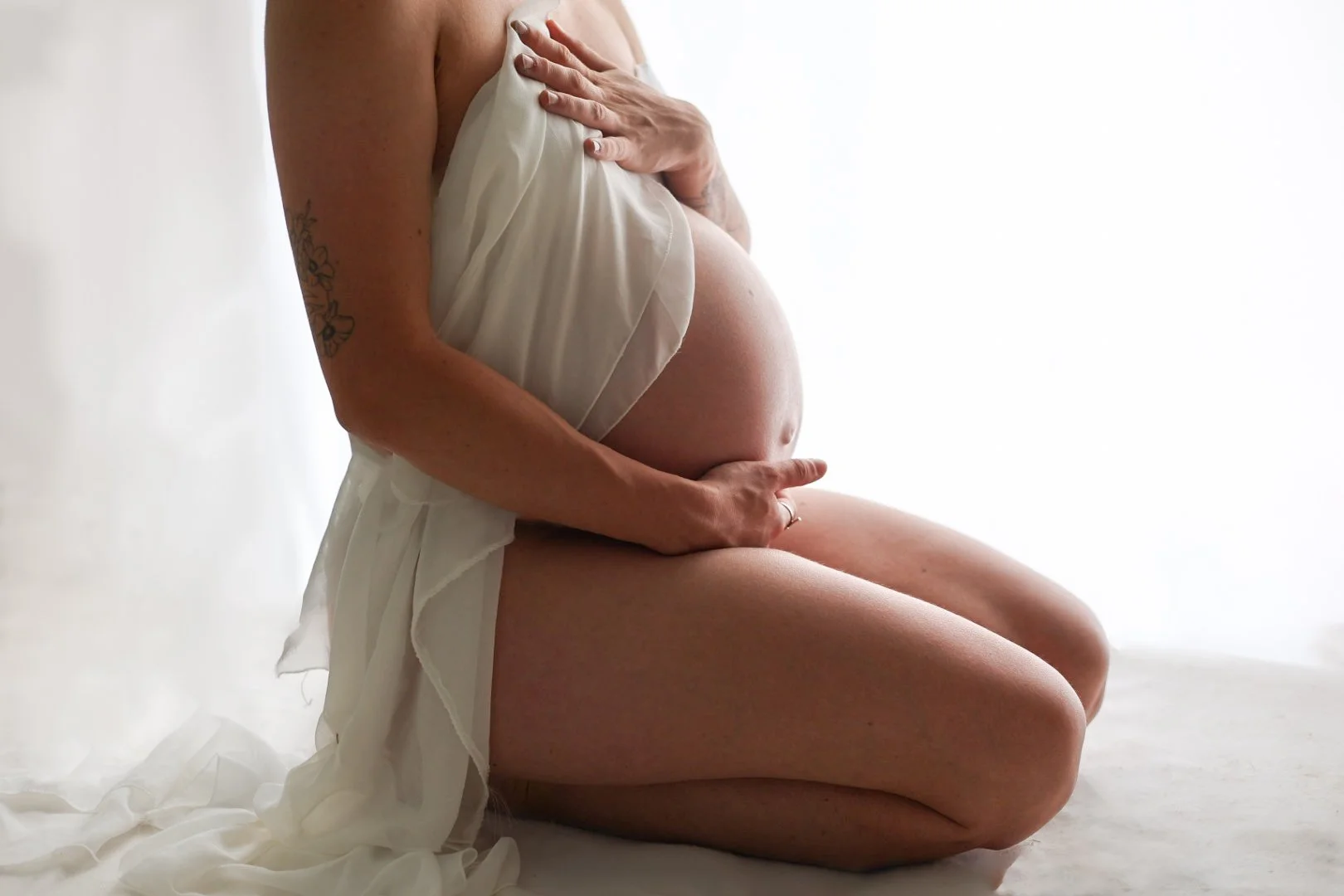 Femme enceinte assise, tenant son ventre avec une main, portant une robe blanche.